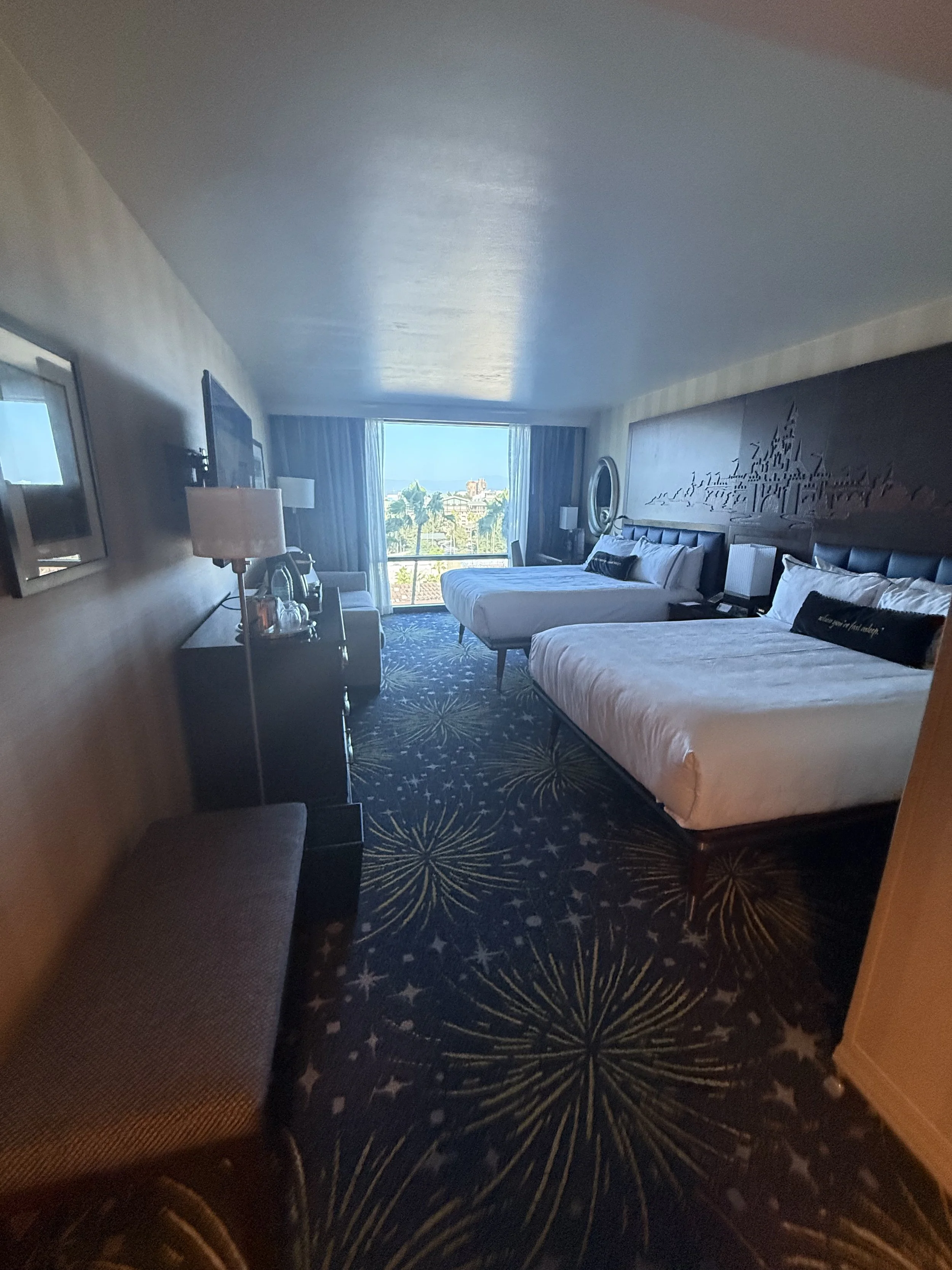 A hotel room with two queen beds, a sitting area, and a large window overlooking trees and buildings in the distance.