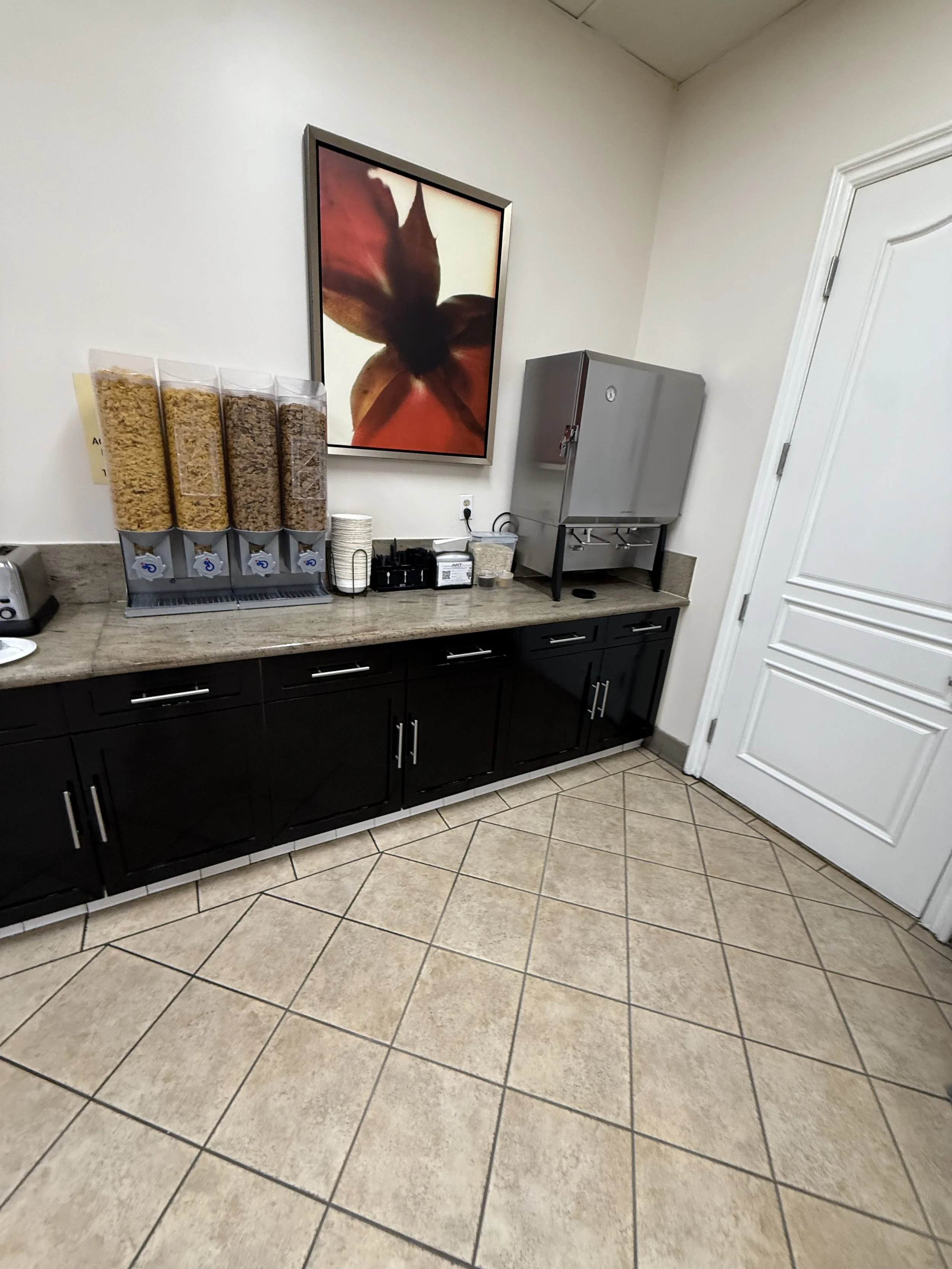 Coffee station with cereal dispensers, cups, and a water boiler, in a room with tiled floor and a framed flower photograph on the wall.