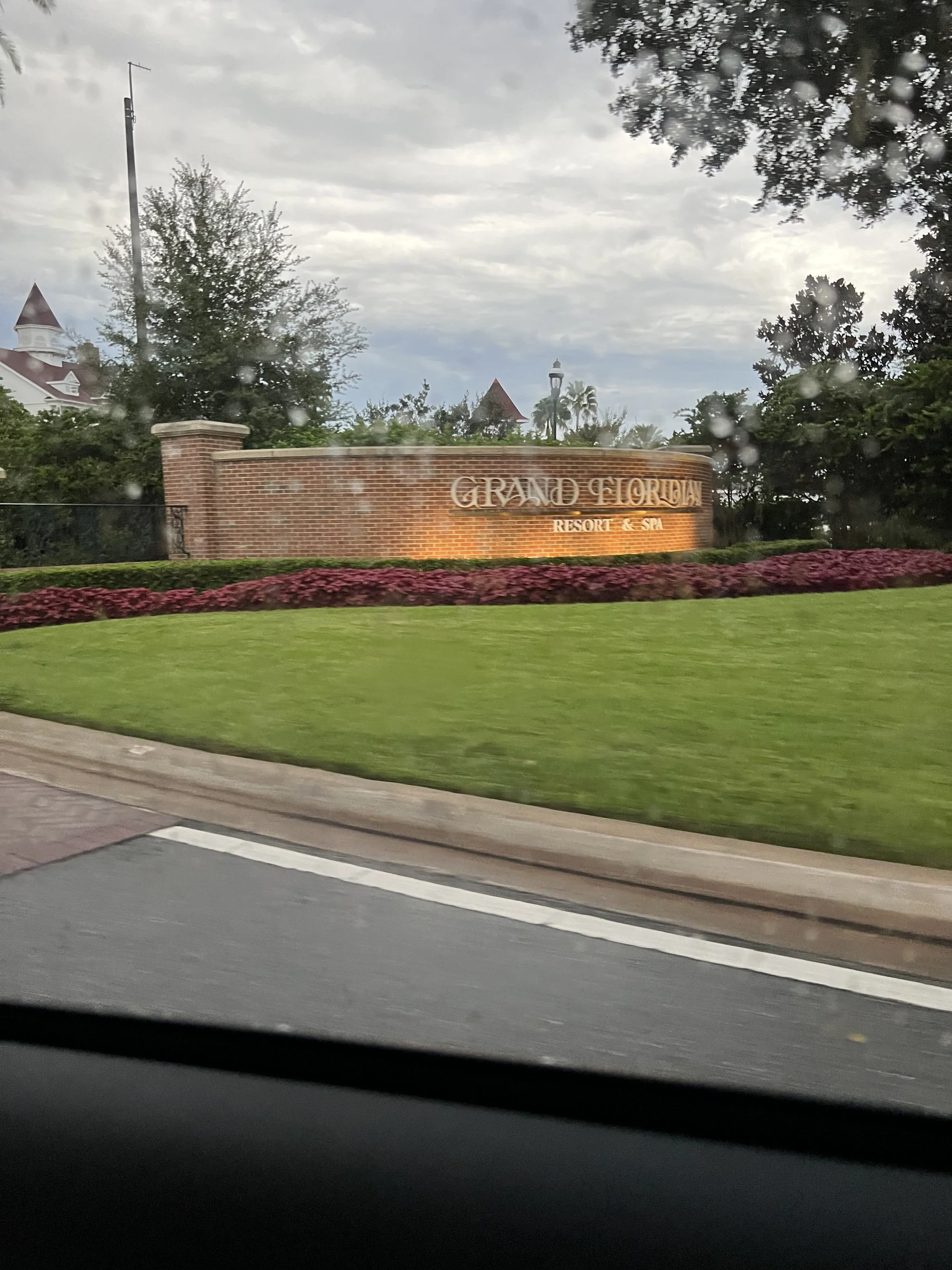 Photo taken from inside a car showing a brick sign for Grand Floridian Resort & Spa, surrounded by greenery and flowers, with a cloudy sky above.