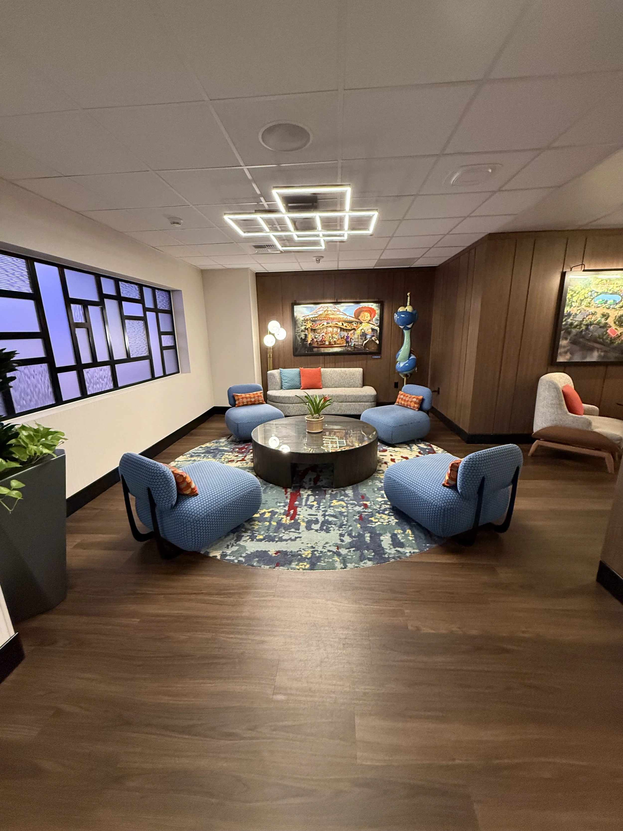 A modern seating area with a circular rug, four blue chairs, a beige sofa, and colorful decor including artwork, lamps, and sculptures.