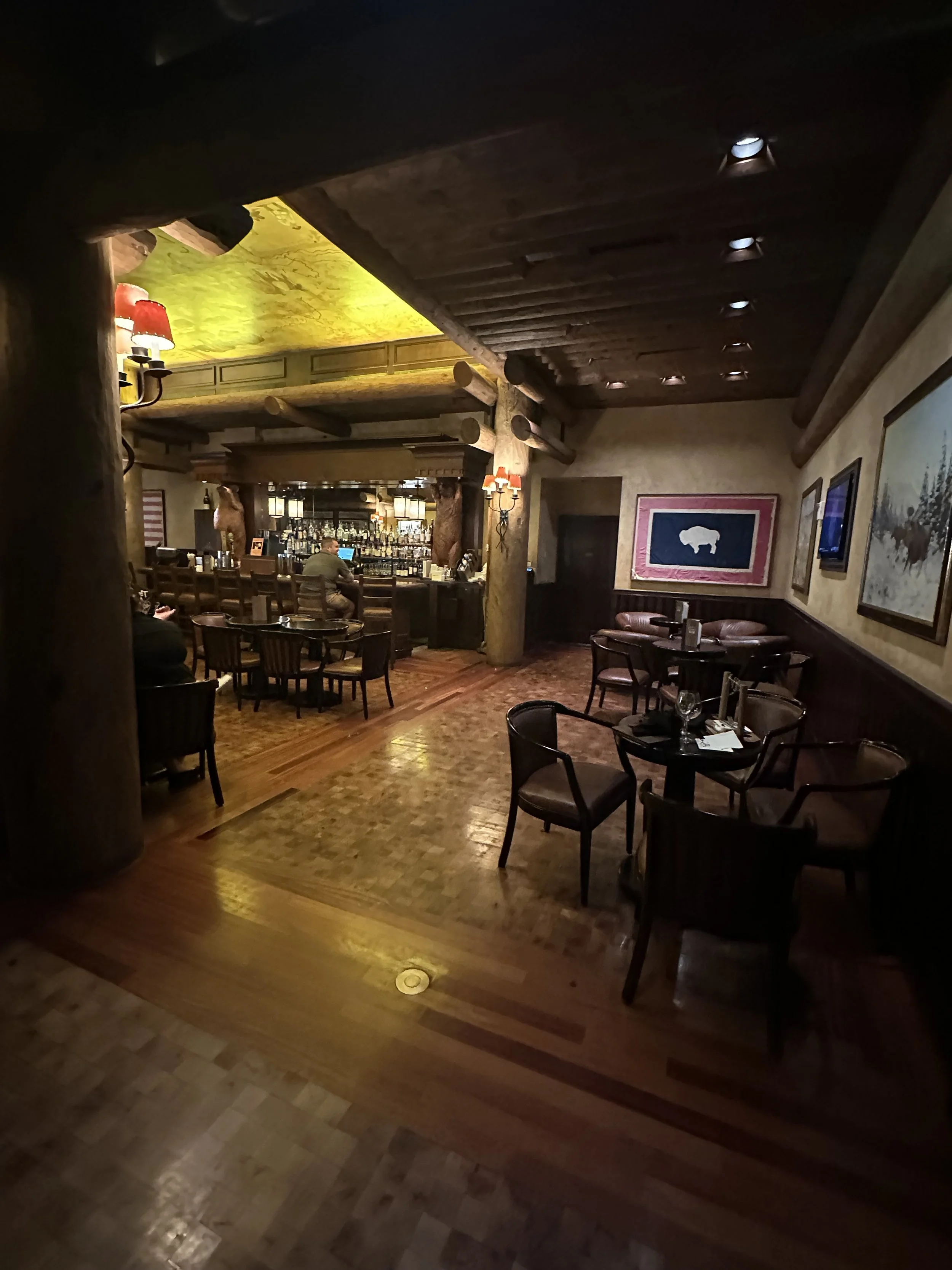 Interior of a cozy, rustic restaurant with wooden floors, dark wood furniture, and softly lit walls decorated with artwork. A bar area with stools is visible in the background.