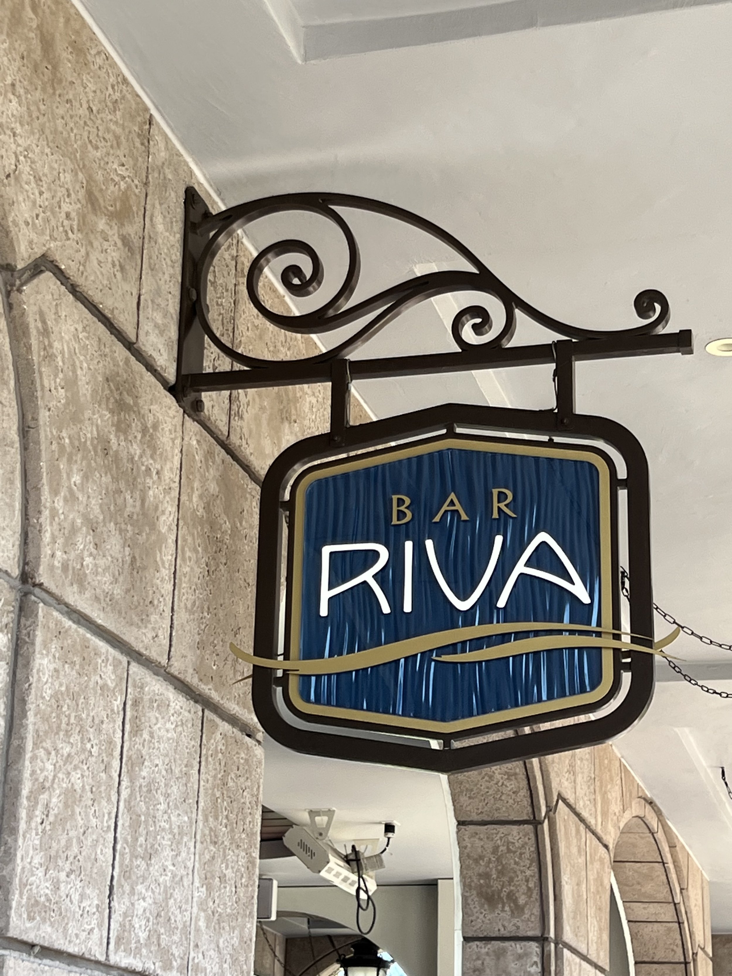 A hanging sign for Bar Riva with a blue background and white neon text, attached to a black wrought iron bracket mounted on a stone wall.