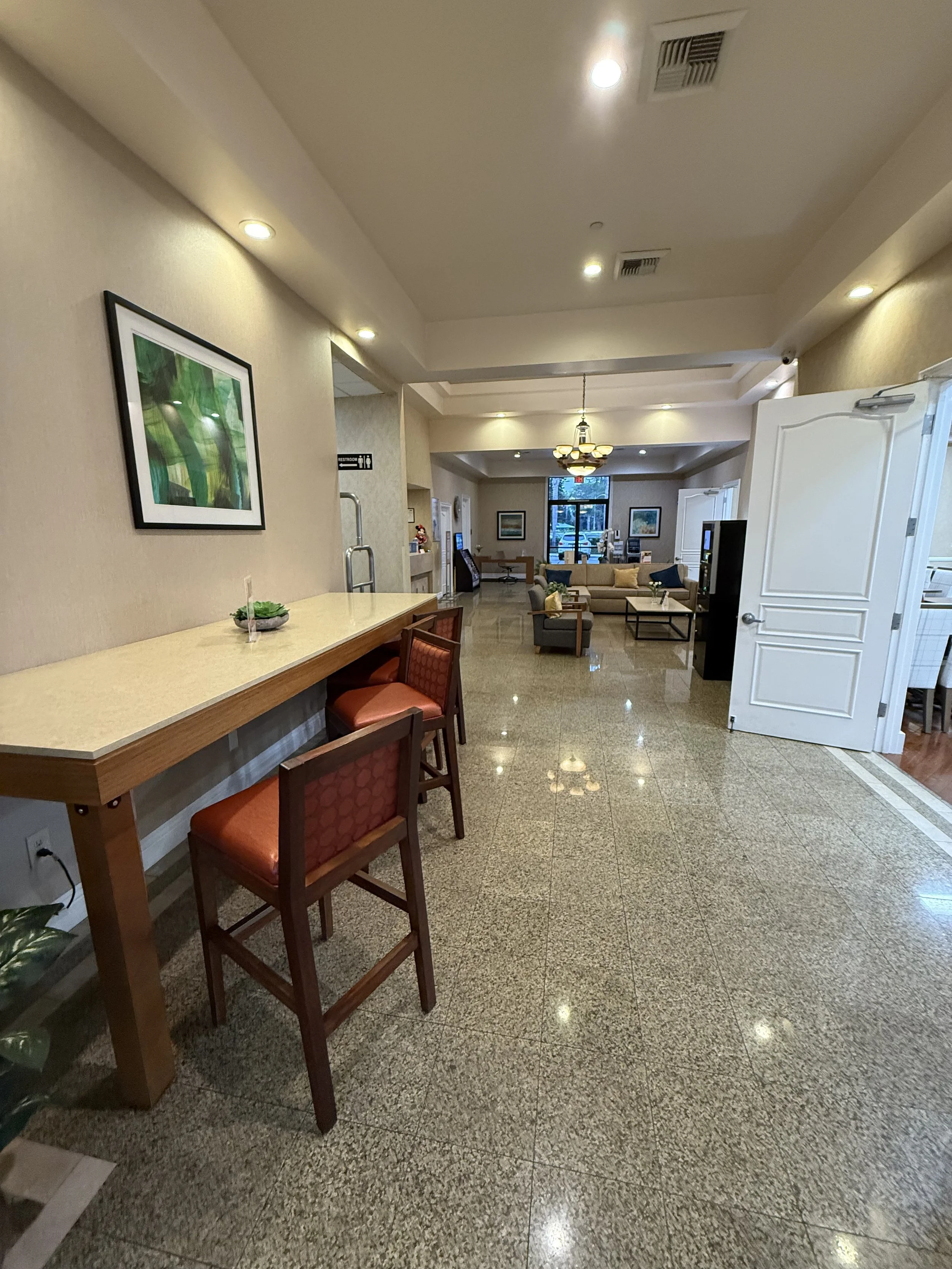 Interior view of a hotel lobby with a high ceiling, a bar-height counter with chairs on one side, a wall-mounted artwork, and seating area with sofas and tables in the background.