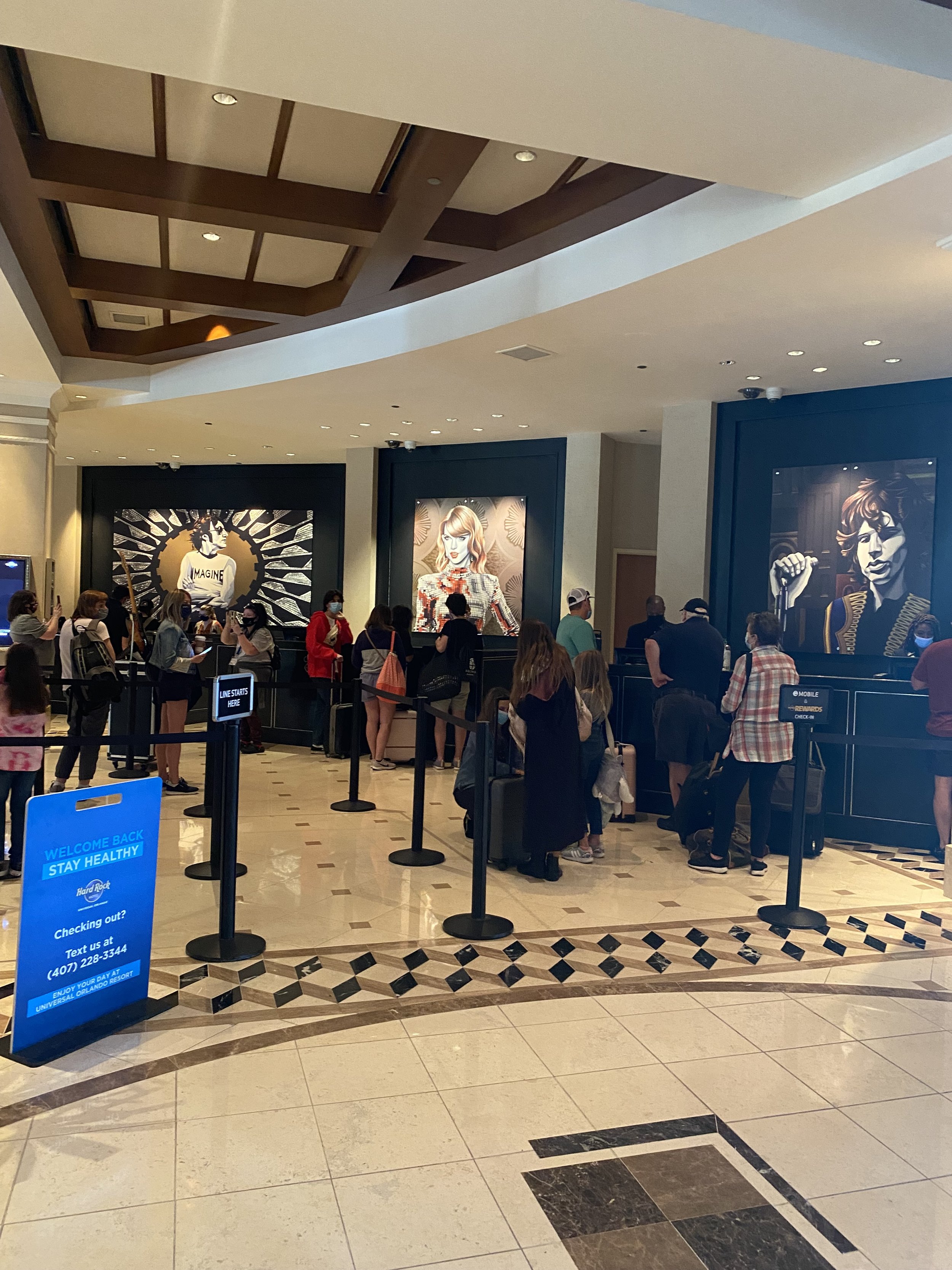 People standing in line at a check-in counter at a hotel or event, with large portraits on the wall behind them and a blue sign that says 'Welcome Back Stay Healthy' in front.