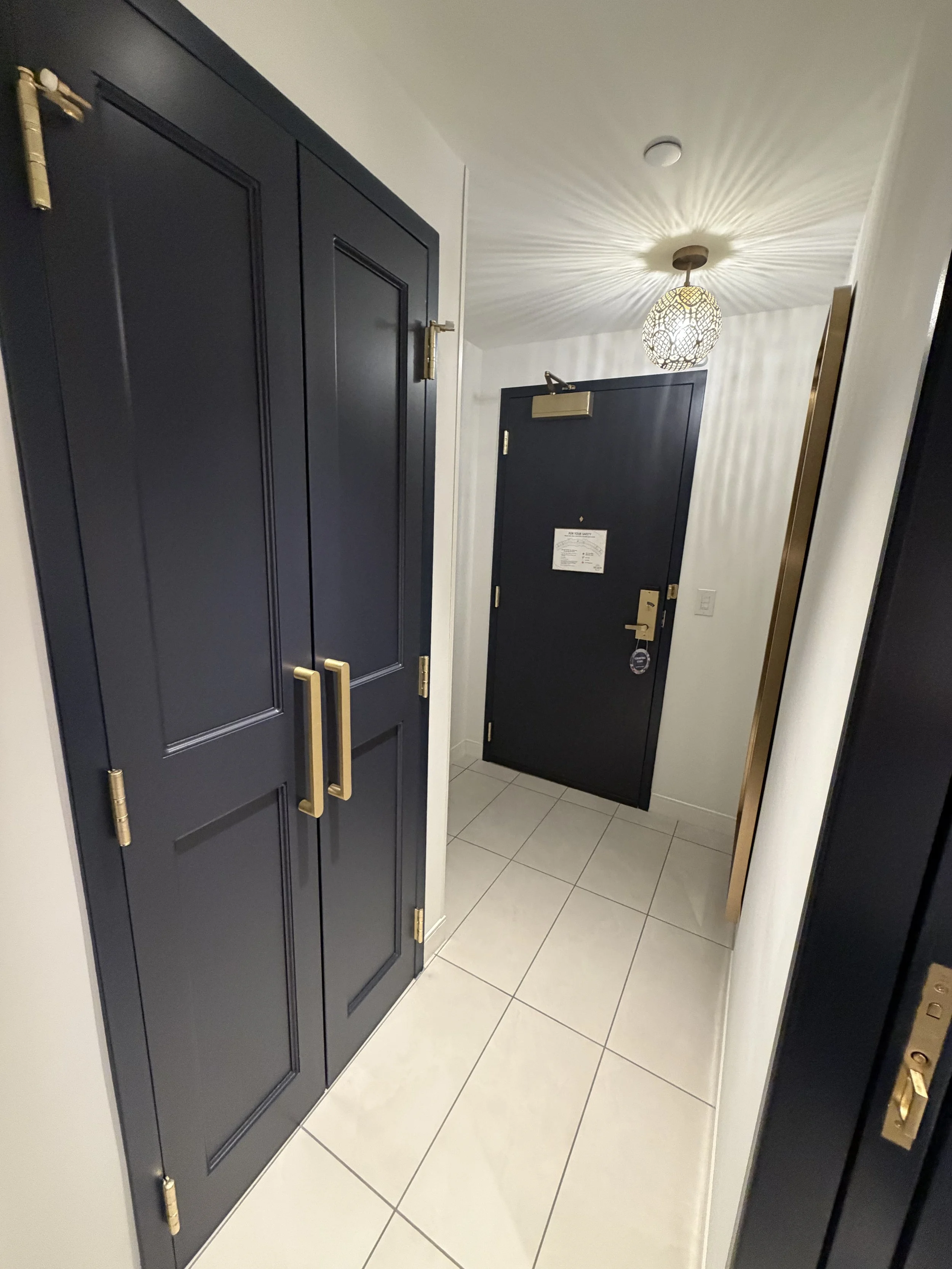 A hallway with dark navy blue doors, gold handles, white tiled floor, and a ceiling light fixture creating a starburst shadow pattern.