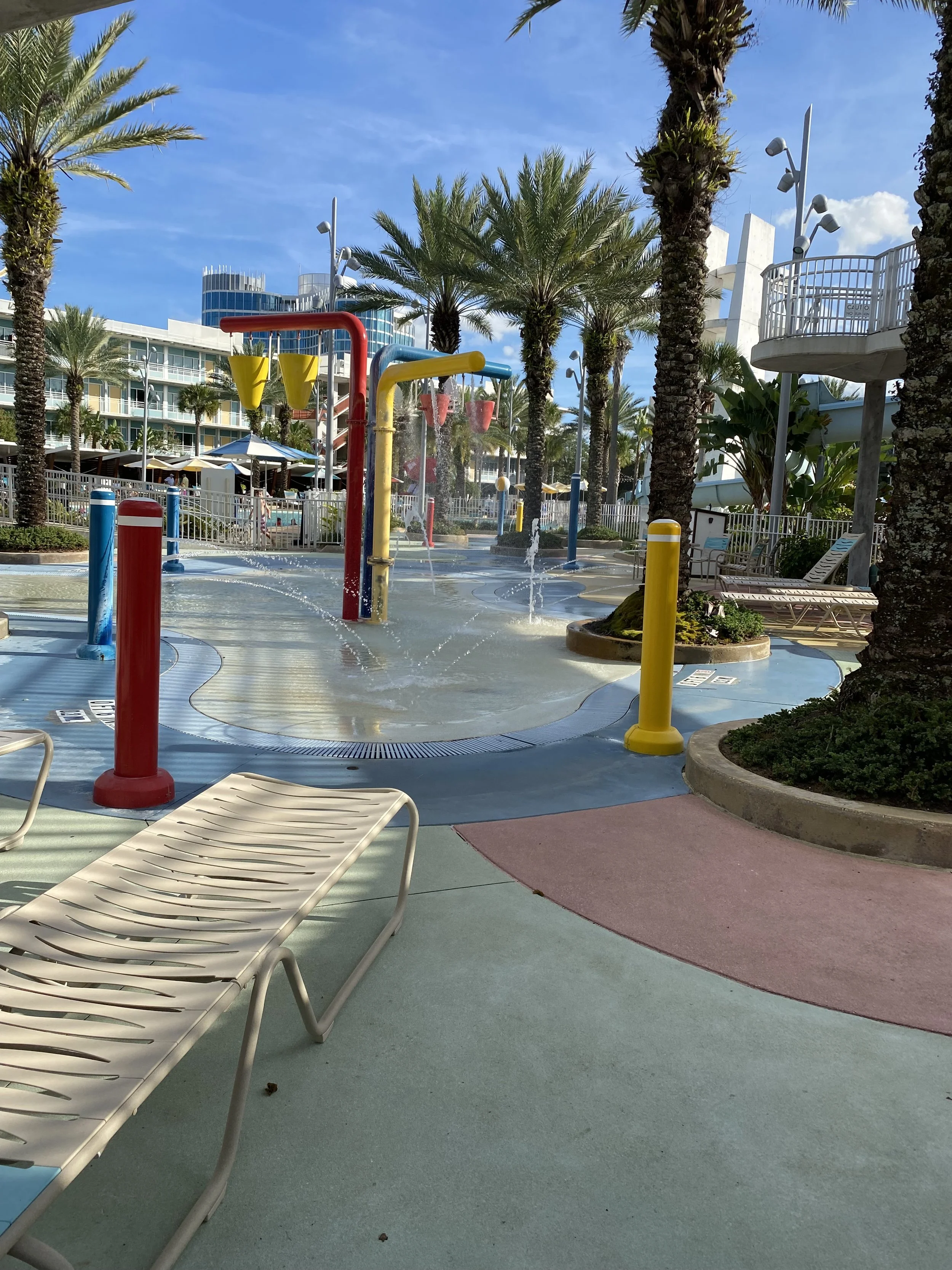 Splash pad with water sprays, surrounded by palm trees, lounge chairs, and colorful posts, with a modern building and blue sky in the background.