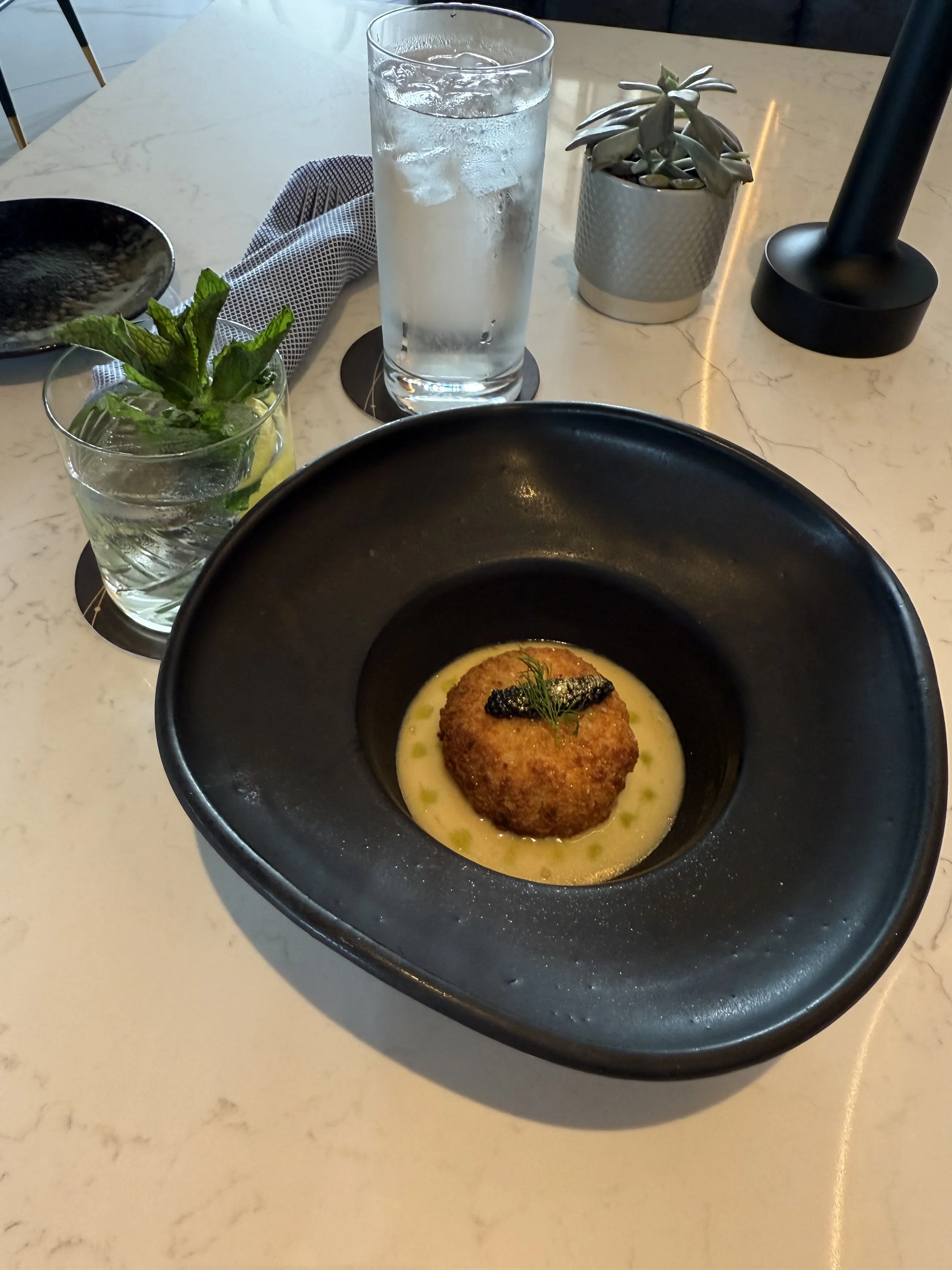 A black bowl with a croquette garnished with caviar, dill, and accompanied by a light brothy sauce, on a white marble table with drinks and a small potted succulent plant.
