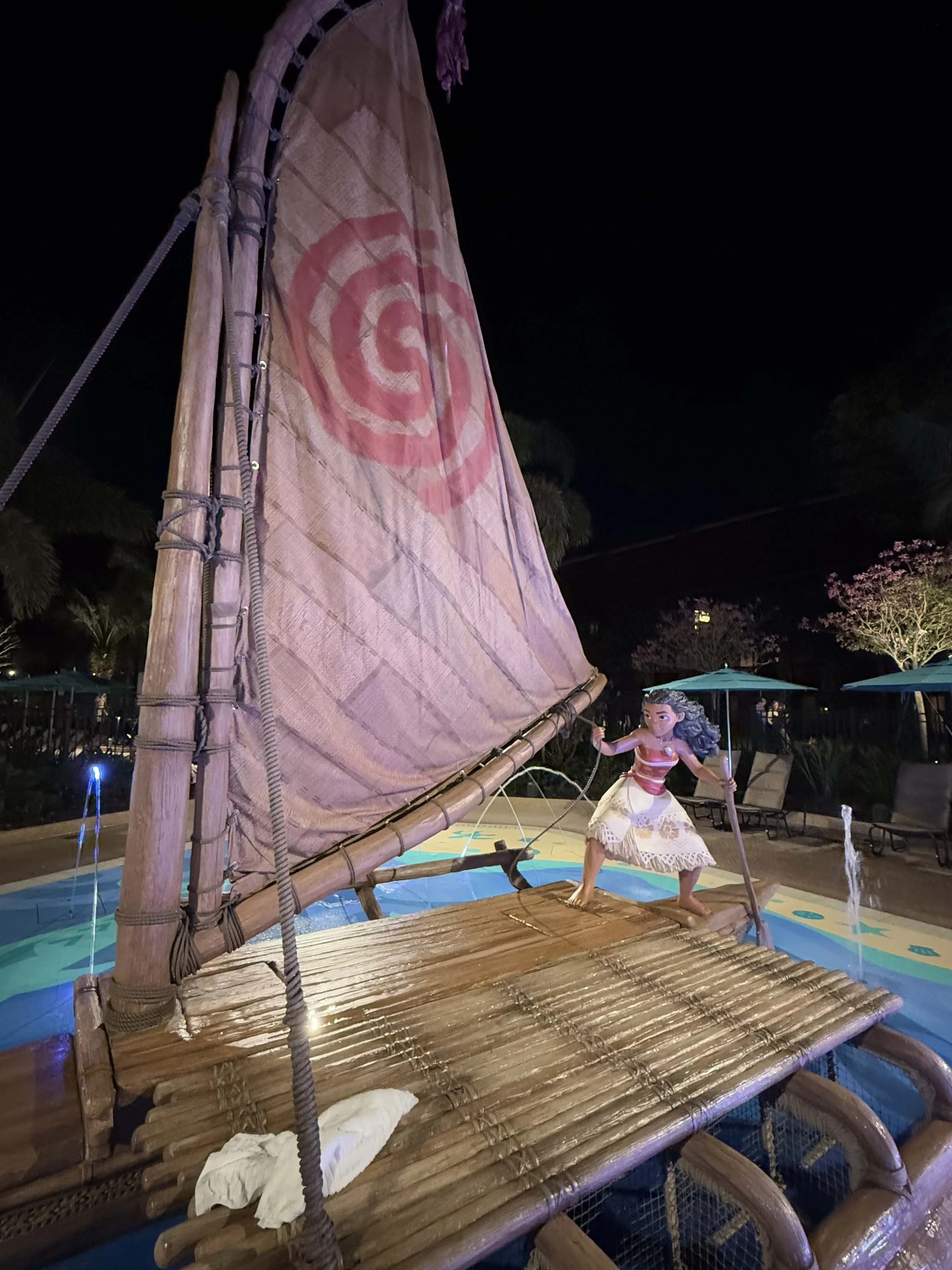 Statue of Moana, a Disney character, standing on a bamboo raft with a sail, in a pool area at night.