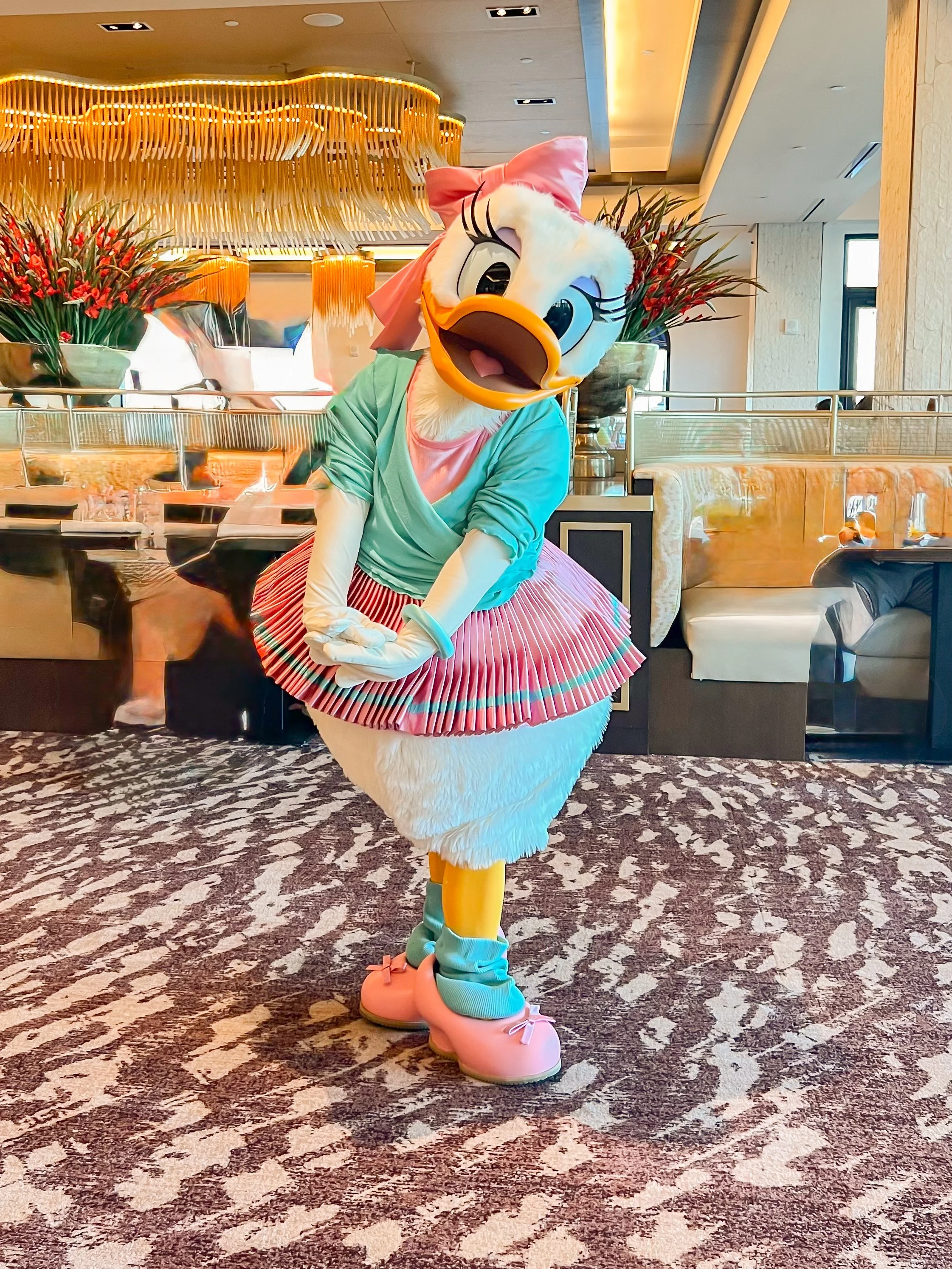 Costumed character of Daisy Duck dressed in colorful clothing standing on patterned carpet in a luxurious hotel lobby with floral arrangements and modern decor.