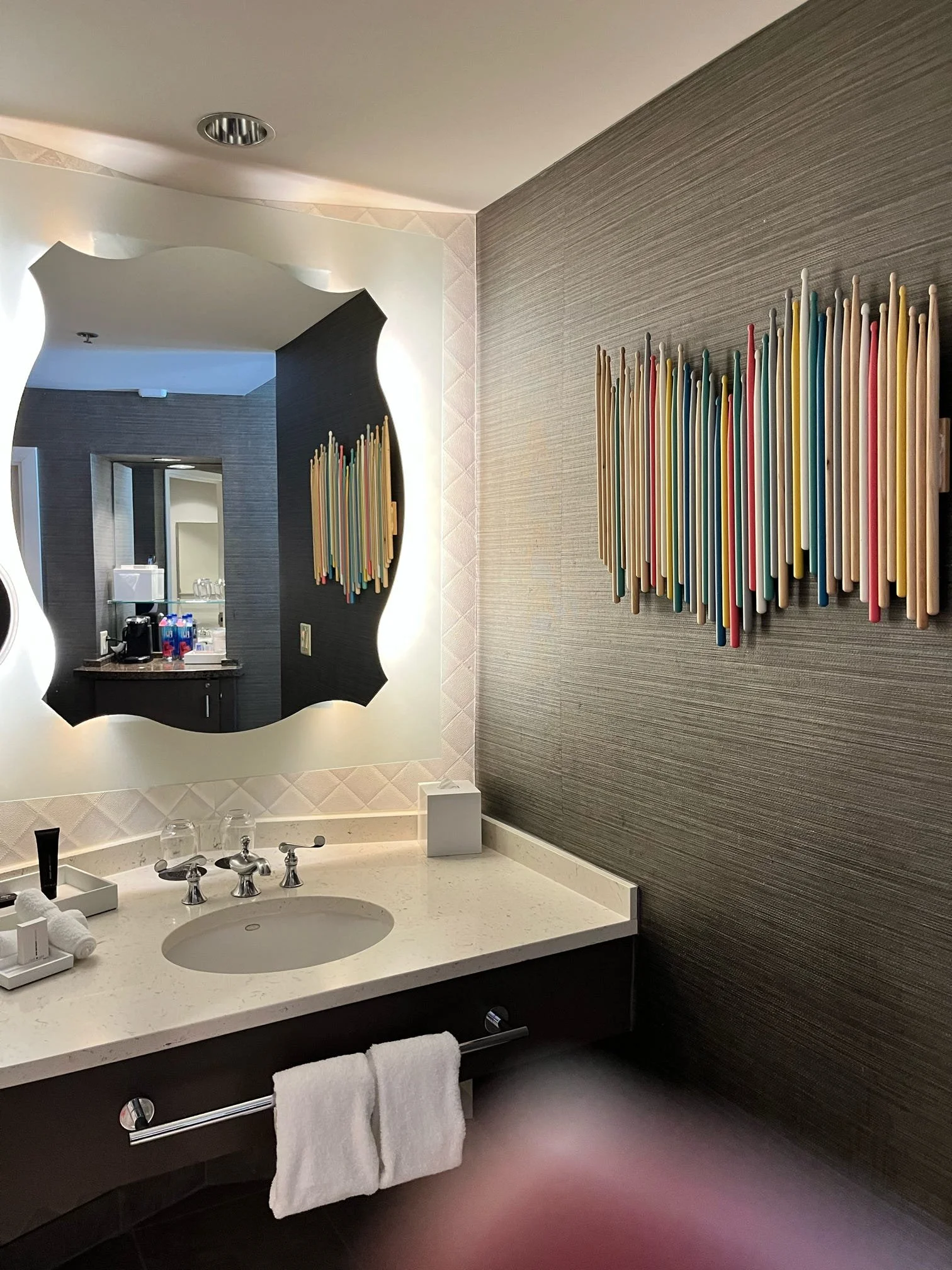 Hotel bathroom sink area with a mirror, colorful wooden art installation on the wall, and a countertop with toiletries.