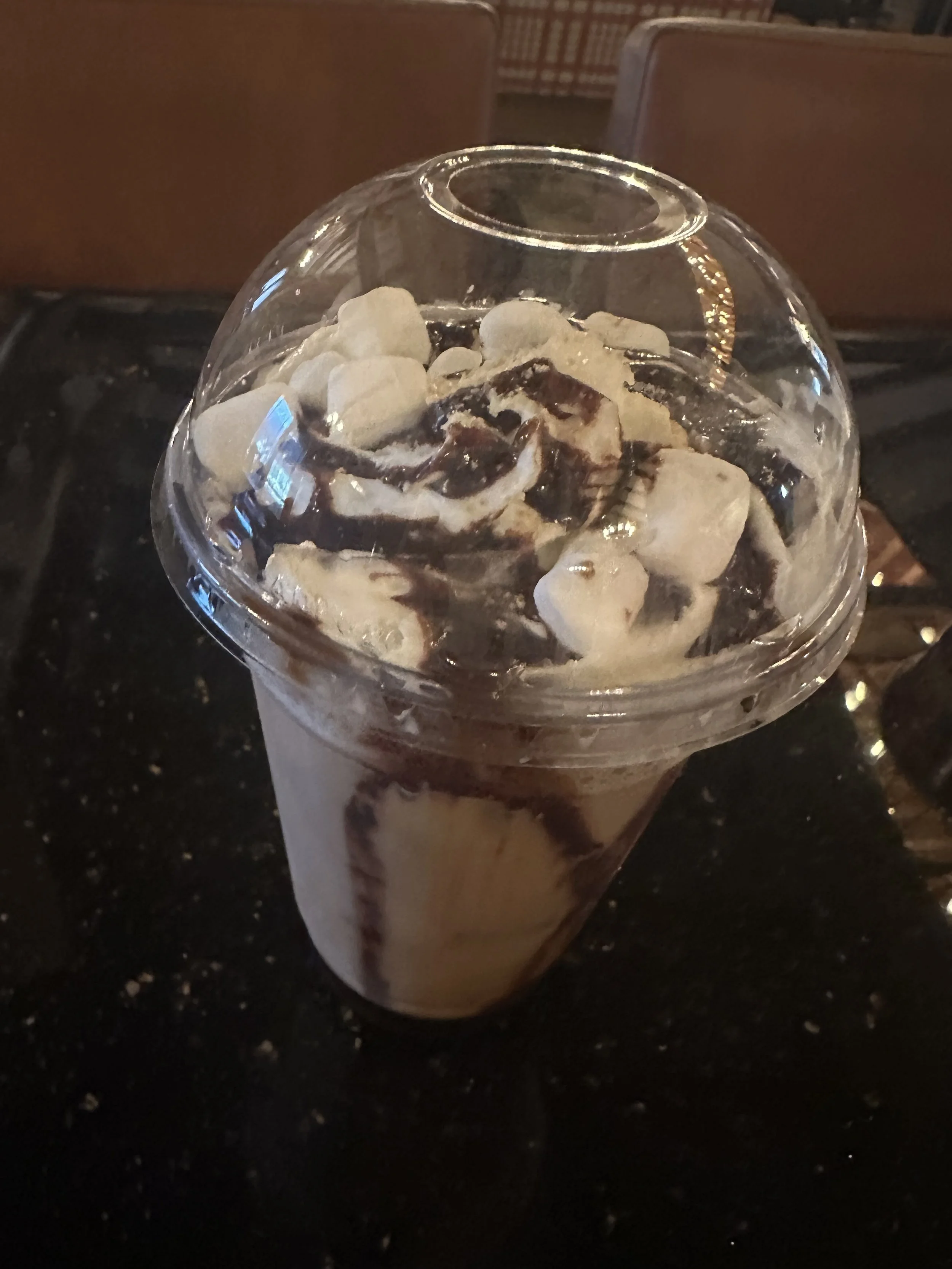 Frappe with whipped cream, chocolate syrup, marshmallows, and a clear plastic dome lid, placed on a dark table in a restaurant setting.
