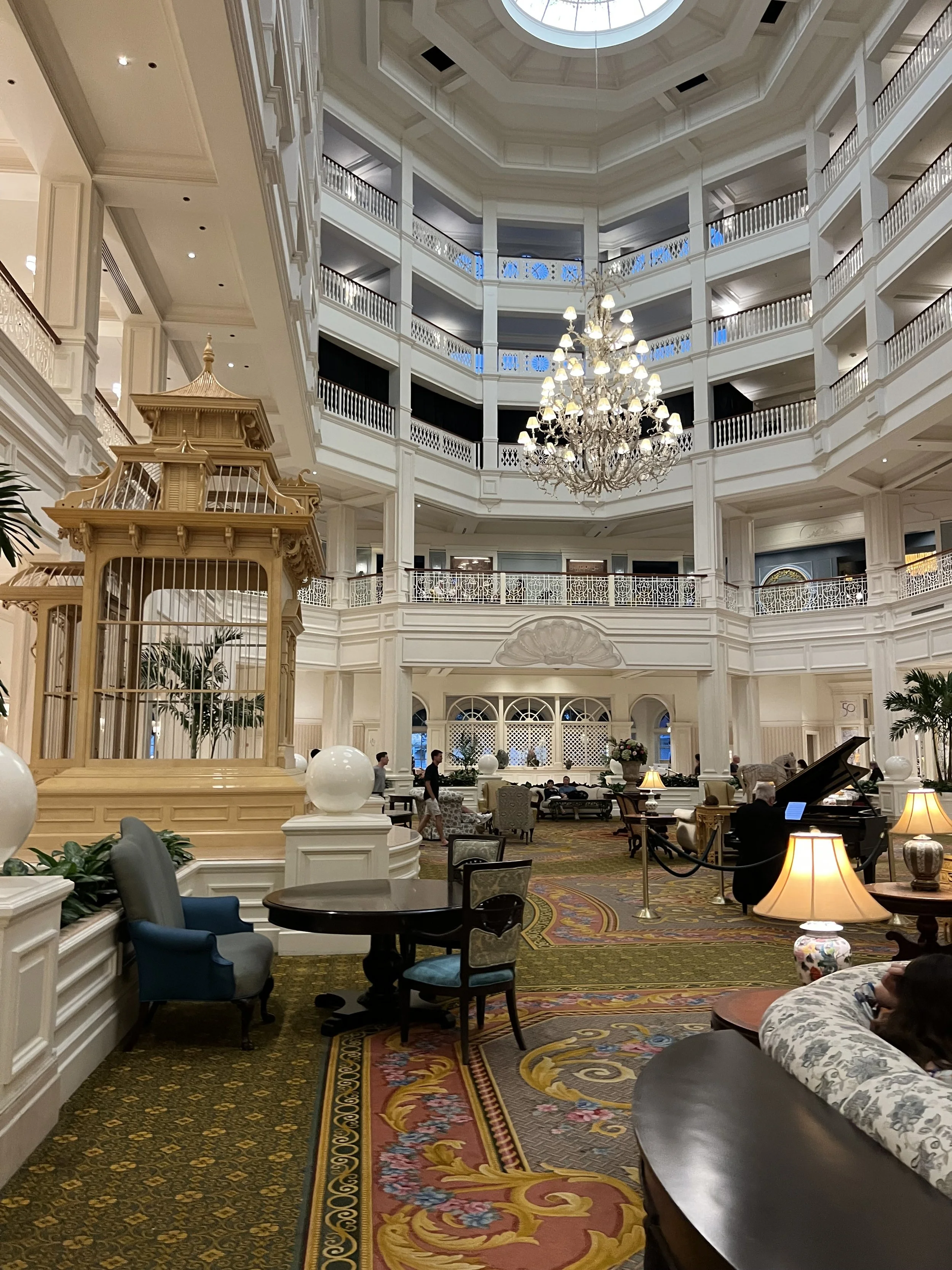 Interior of a luxurious hotel lobby with a high ceiling, a grand chandelier, a large decorative birdcage, and various seating areas, including a person playing the piano.