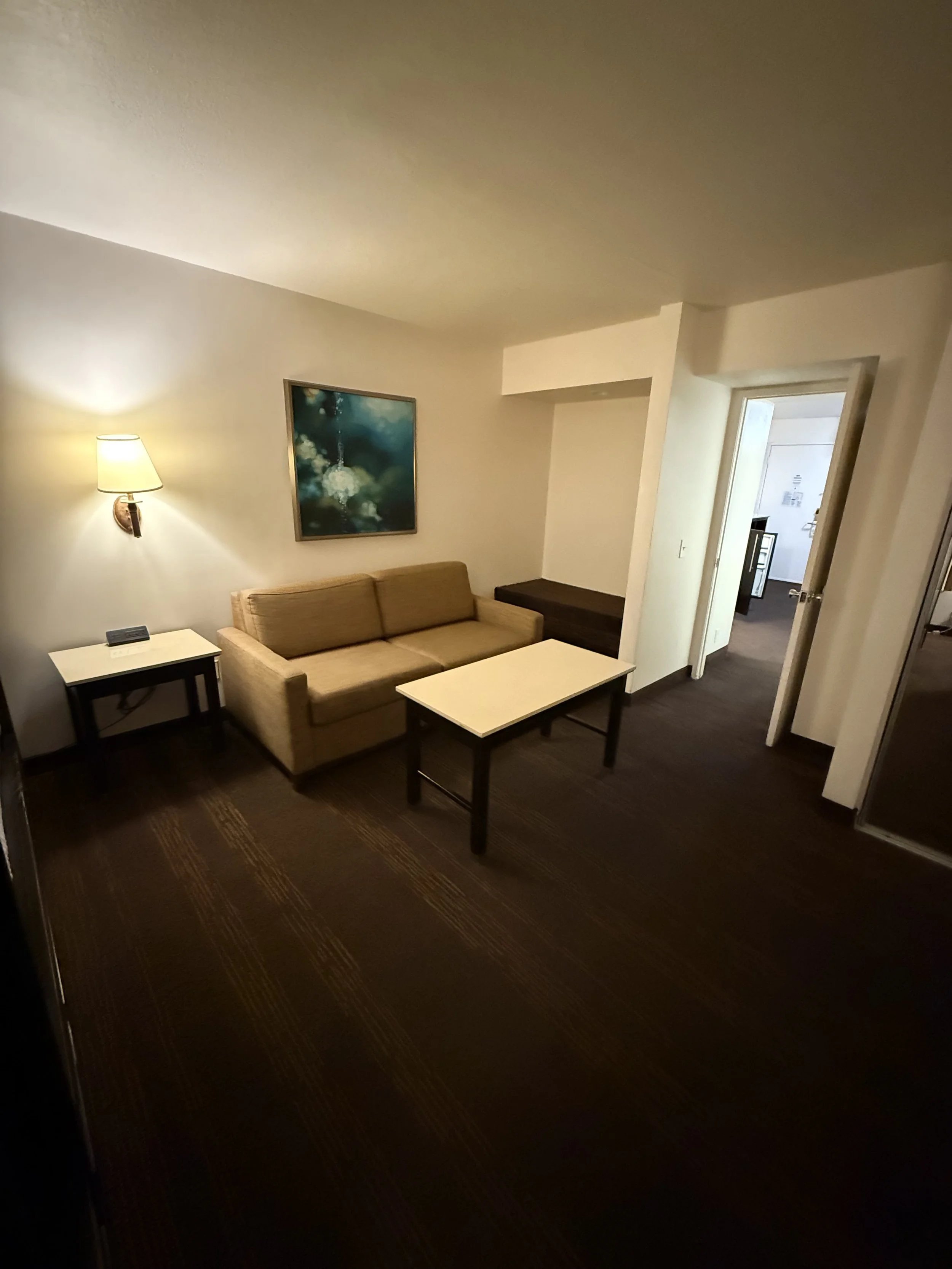 Hotel room with beige sofa, dark wood coffee table, side table, wall art, lamp, and doorway to another room.