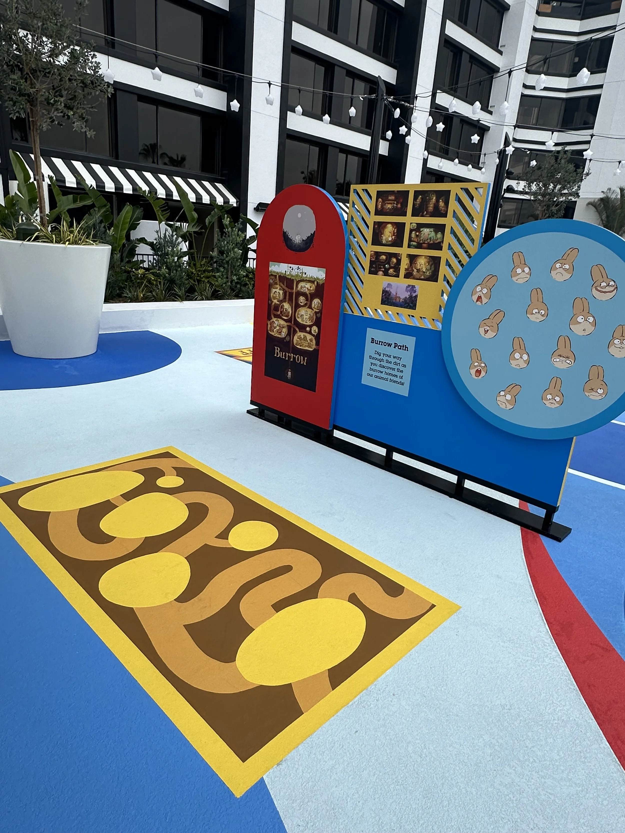 Colorful outdoor art installation with cartoon bunny faces, a red sign with pictures and a sign with the word 'Burrow', and a blue panel with bunnies, on a blue and red painted ground, next to a large white planter, in front of a modern building.