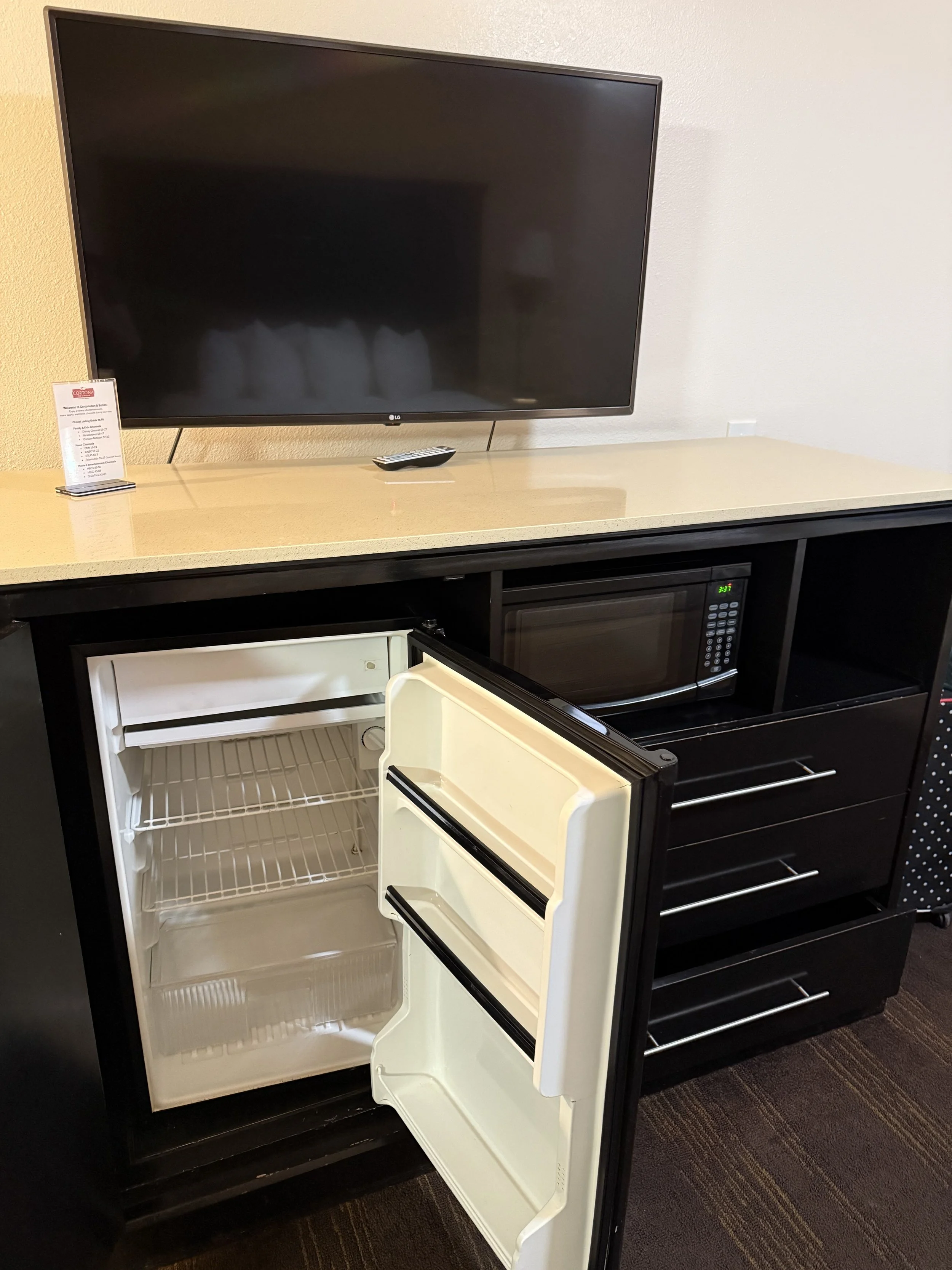 Small open refrigerator with empty shelves below a countertop, microwave oven, and a large flat-screen TV on the wall above.
