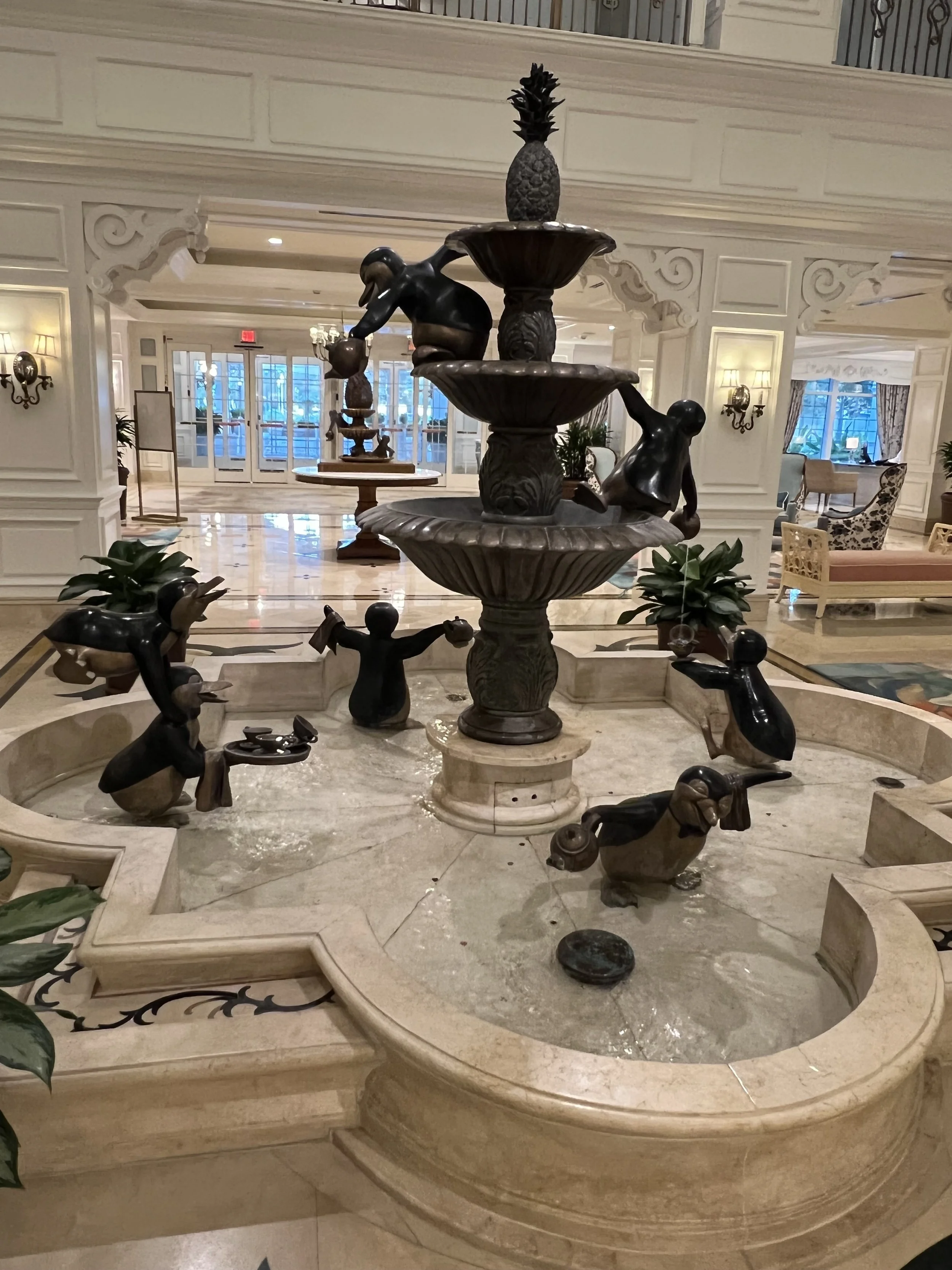 Indoor marble fountain with bronze sculptures of children playing, featuring a three-tiered fountain, in a luxurious hotel lobby with ornate white paneling, plants, and seating area in the background.