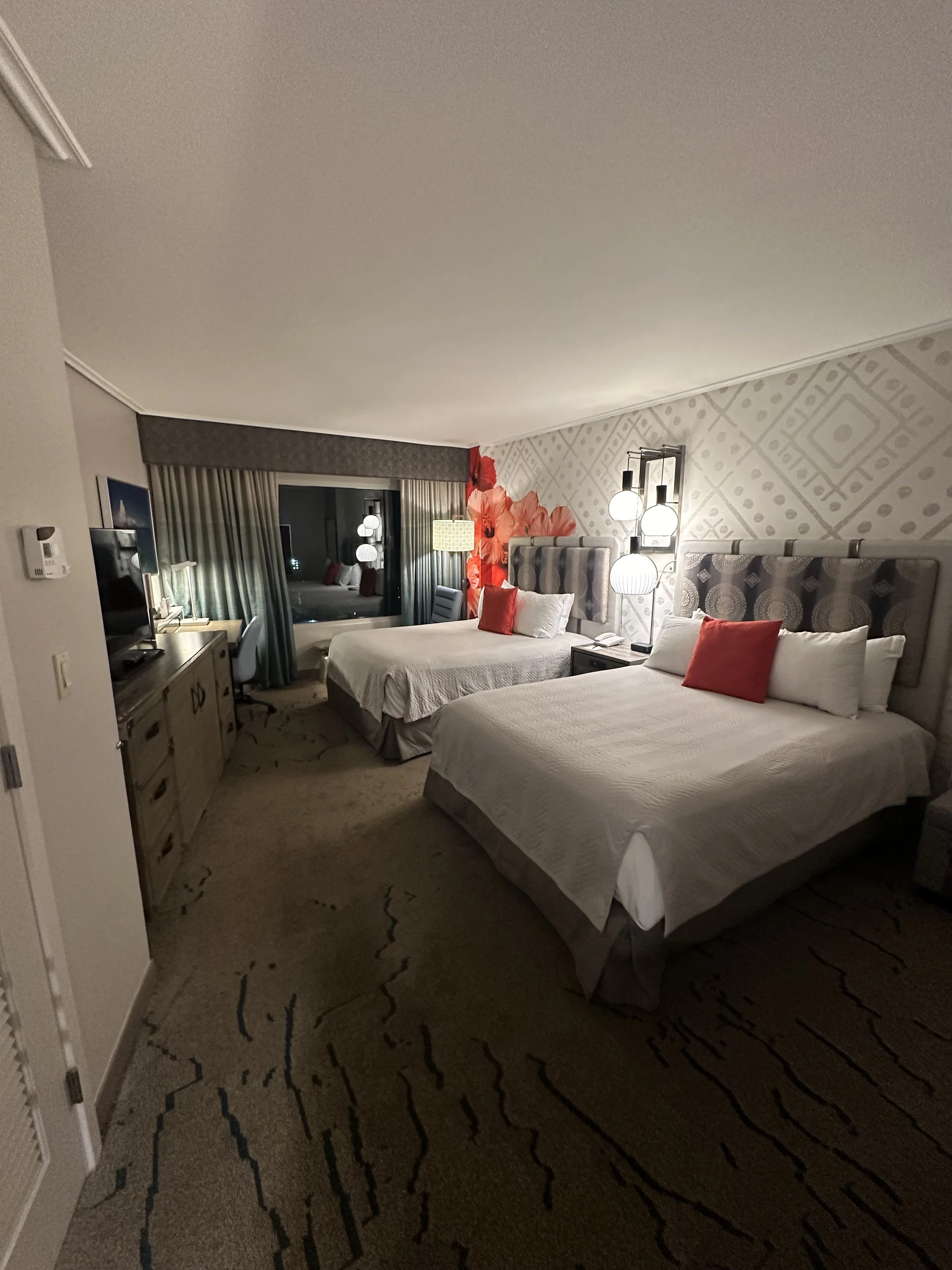A hotel room with two queen beds, each with red pillows, floral headboards, a window with curtains, and a desk with a chair, a TV, and lamps.