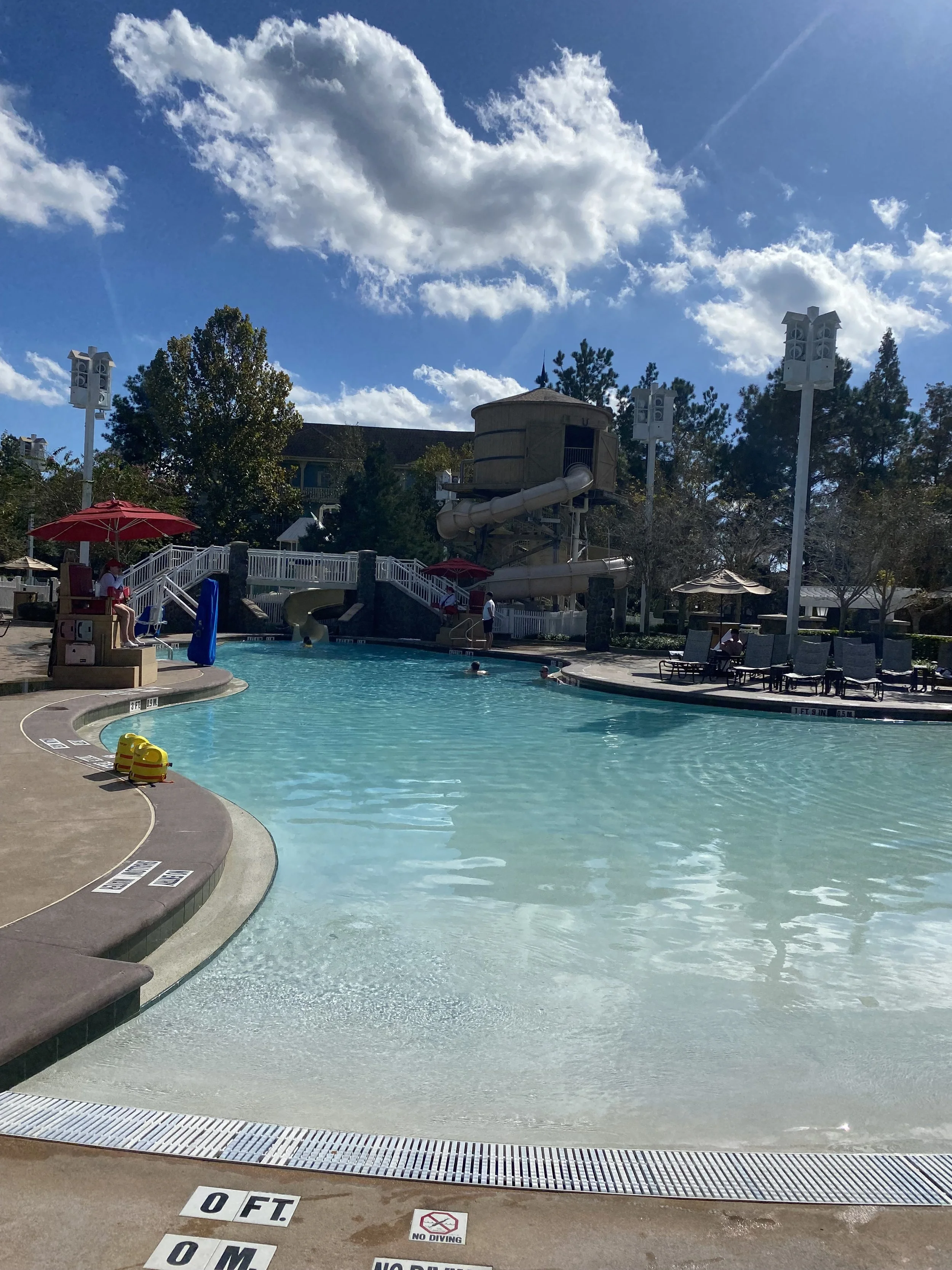 A swimming pool area with a water slide structure in the background, lounge chairs, umbrellas, and a few people swimming and relaxing by the pool on a sunny day with a blue sky and scattered clouds.