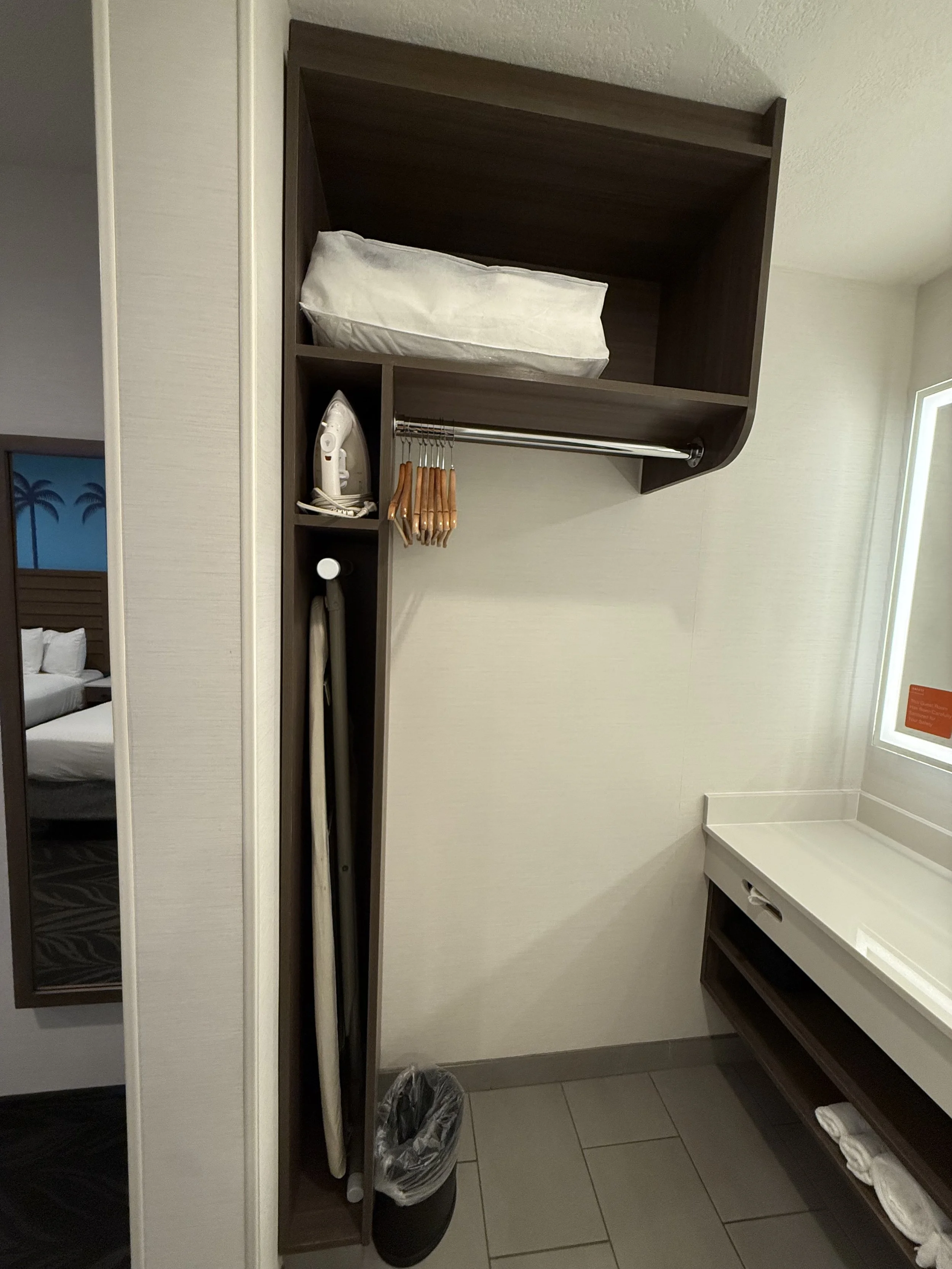 Hotel room closet with a pillow on the top shelf, an iron, a few hangers, an ironing board, and a trash can.