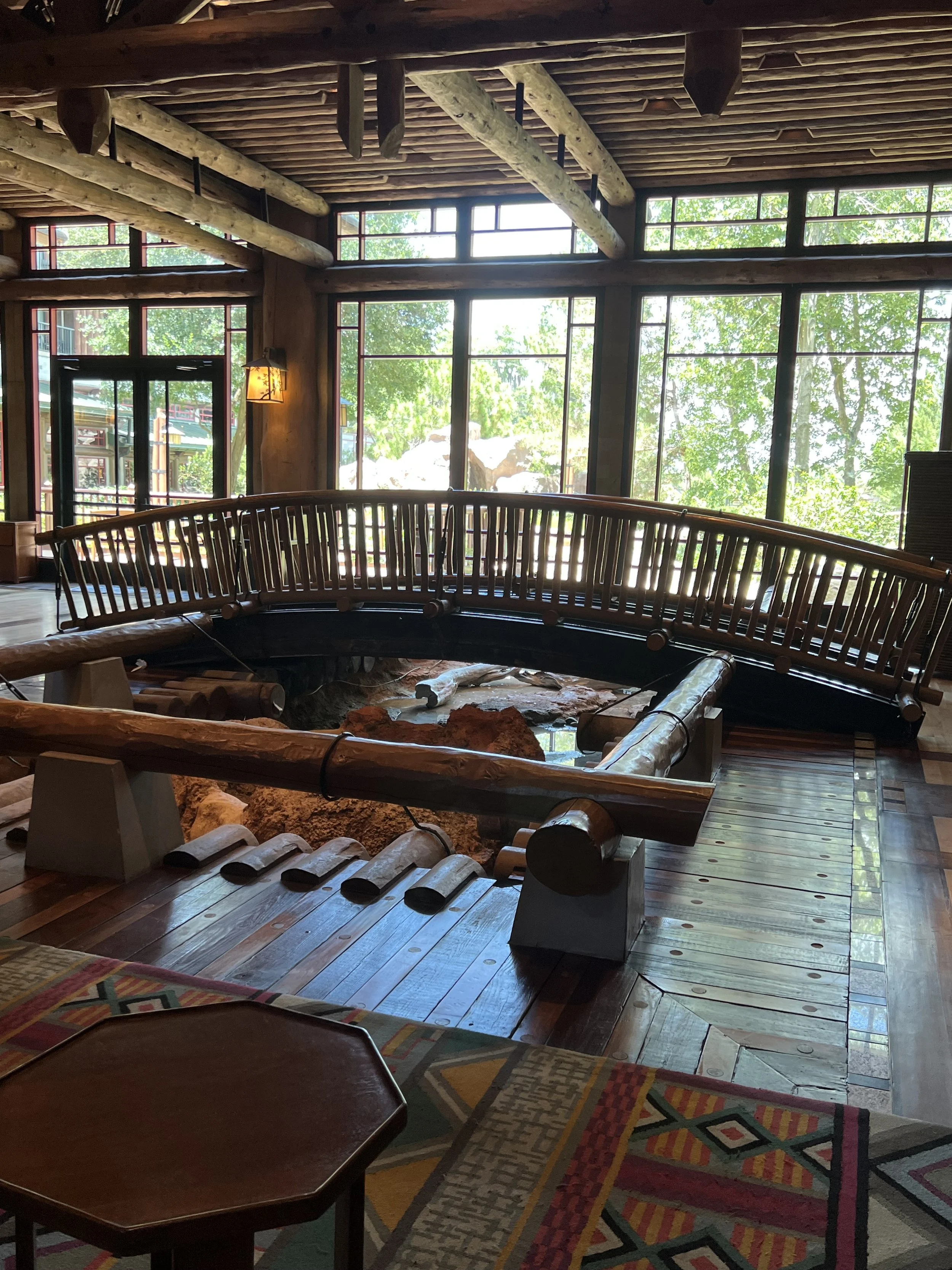 Indoor scene with large windows, wooden logs, and a hot spring pool surrounded by a wooden railing, sunlight filtering through the trees outside.