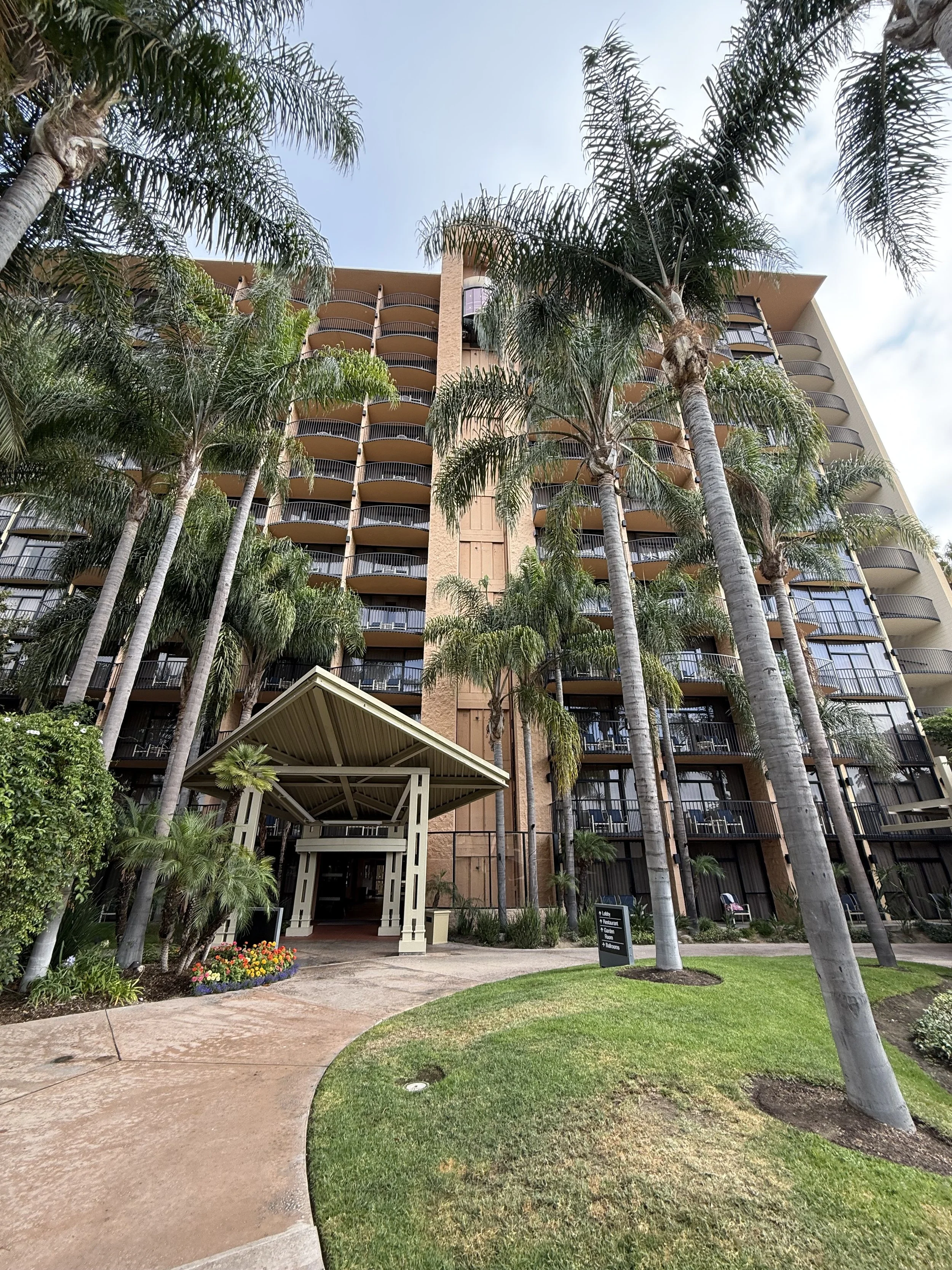 Tall modern apartment building with multiple balconies, surrounded by tall palm trees and a well-maintained grassy area with a curved walkway leading to the entrance. A small garden with colorful flowers is near the walkway, and a sign is visible in 