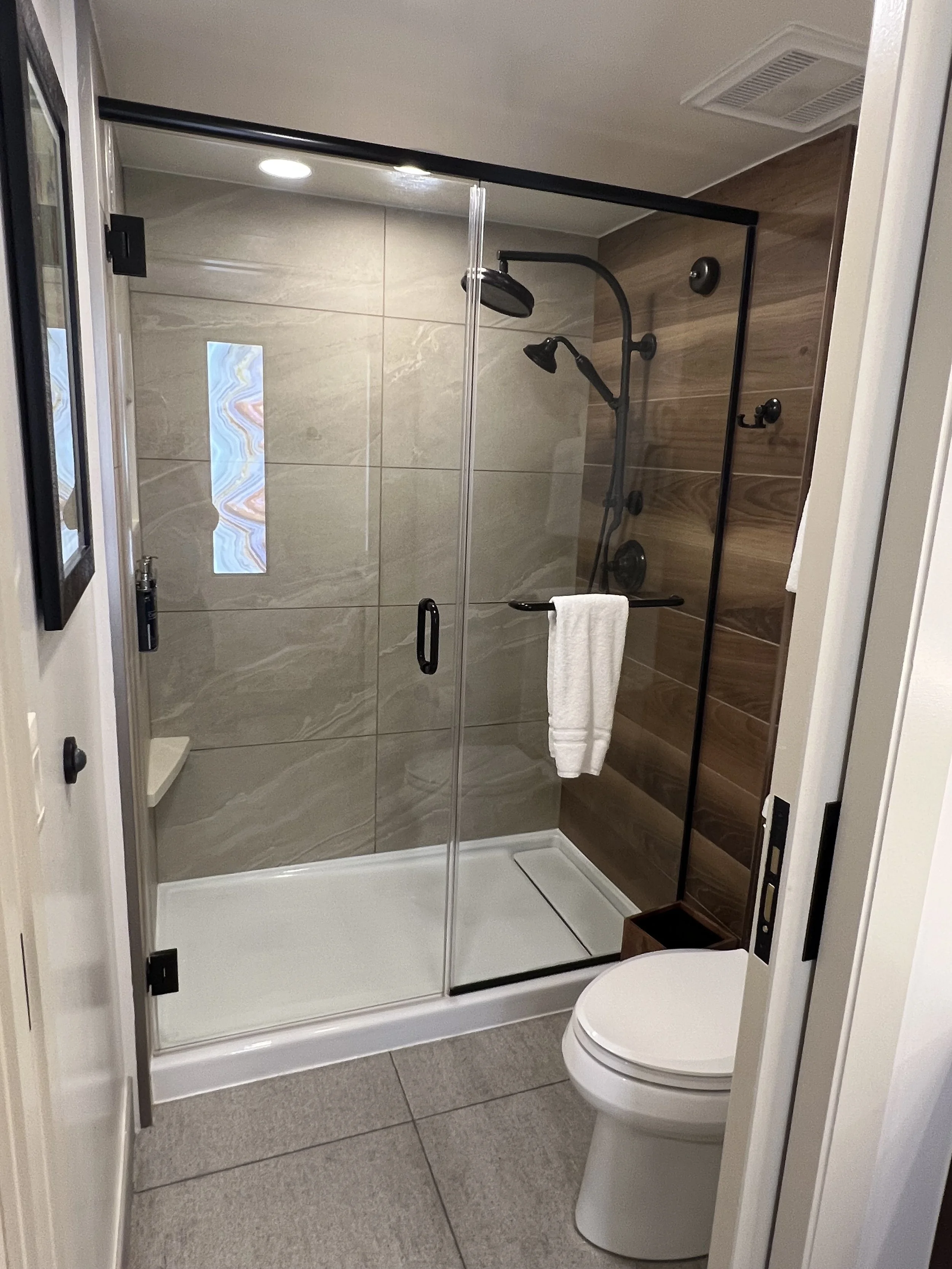 A bathroom with a glass-enclosed shower featuring a rainfall shower head, a hand shower, a towel bar with a white towel, a small window, and wood paneling on one wall.