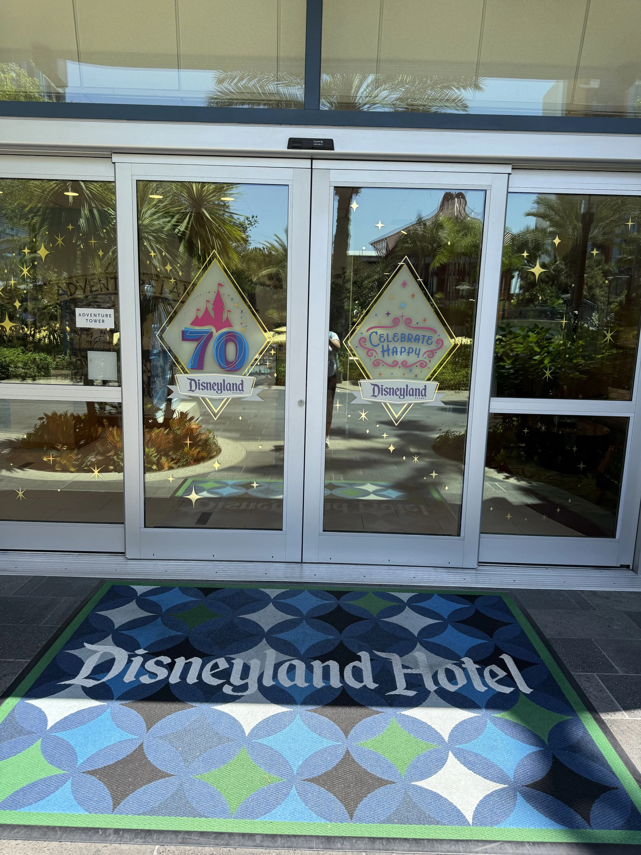 Glass doors with Disneyland decorations celebrating the park's 70th anniversary, featuring colorful signs that read "70 Disneyland" and "Celebrate Happy," with a Disneyland Hotel doormat in front.