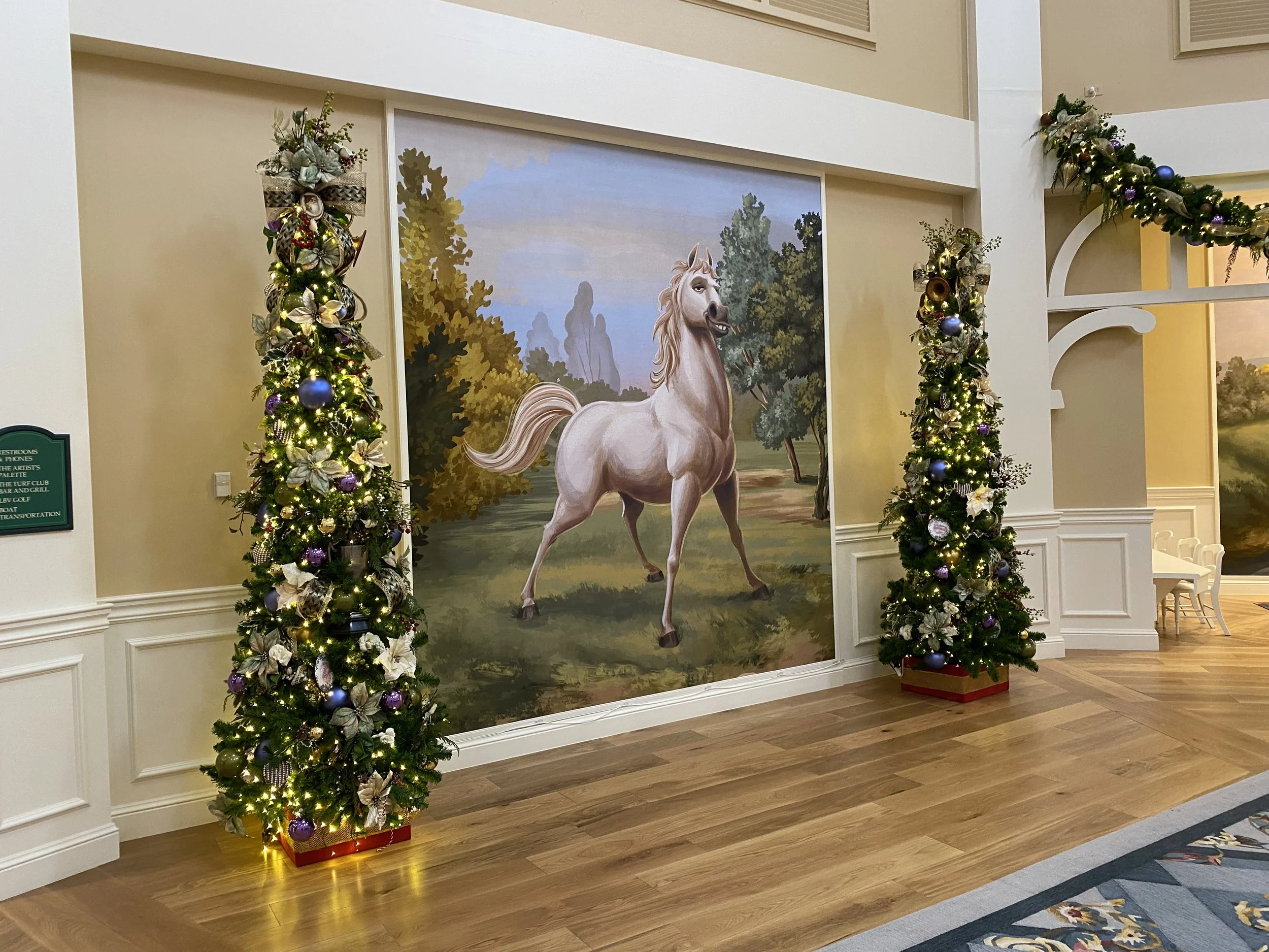 Decorative holiday scene with two decorated Christmas trees and a large painted mural of a white horse on a landscape background.