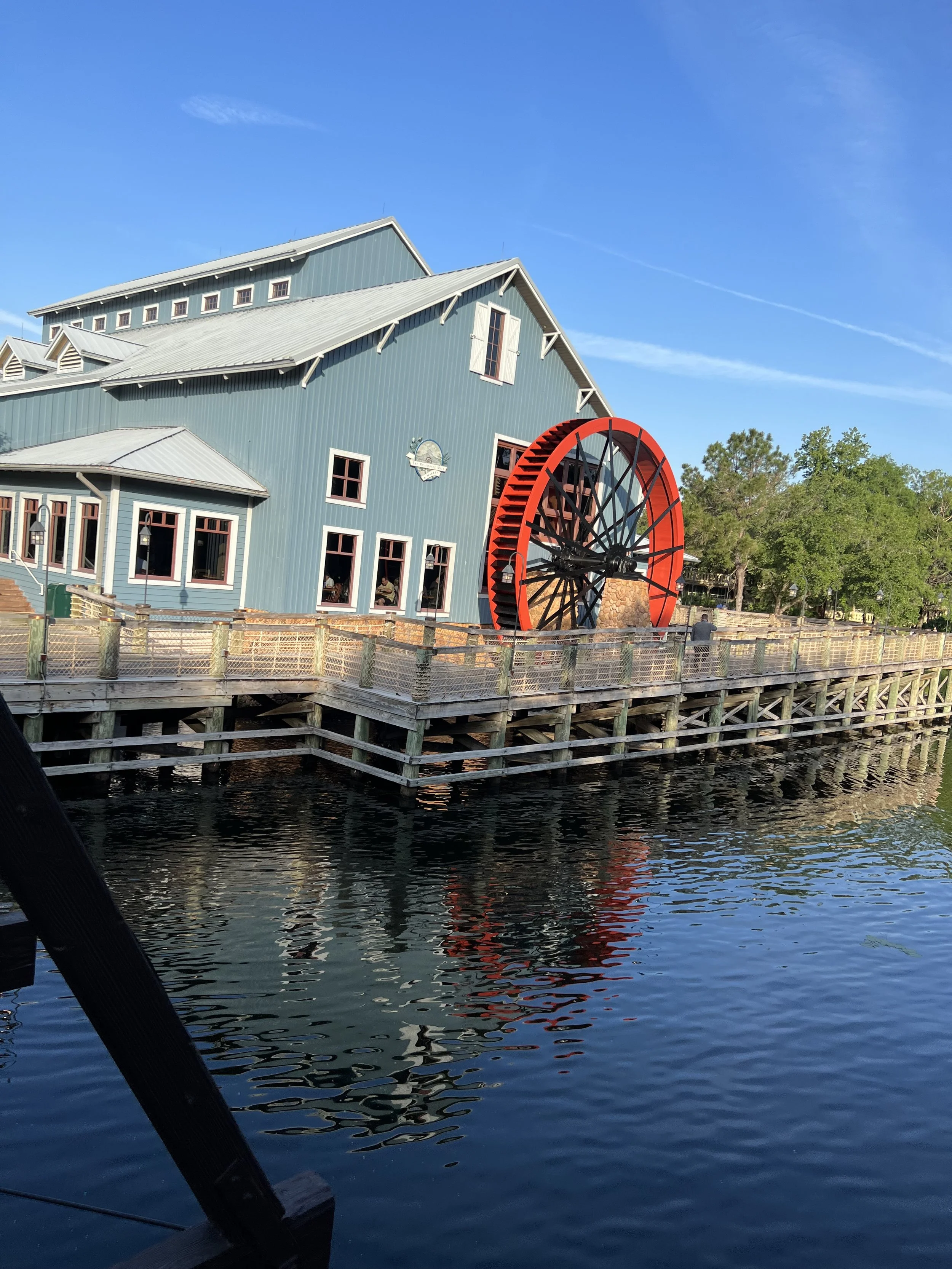 A large historic watermill with a blue building and a red waterwheel situated on a wooden pier over a body of water with trees in the background and a clear blue sky.