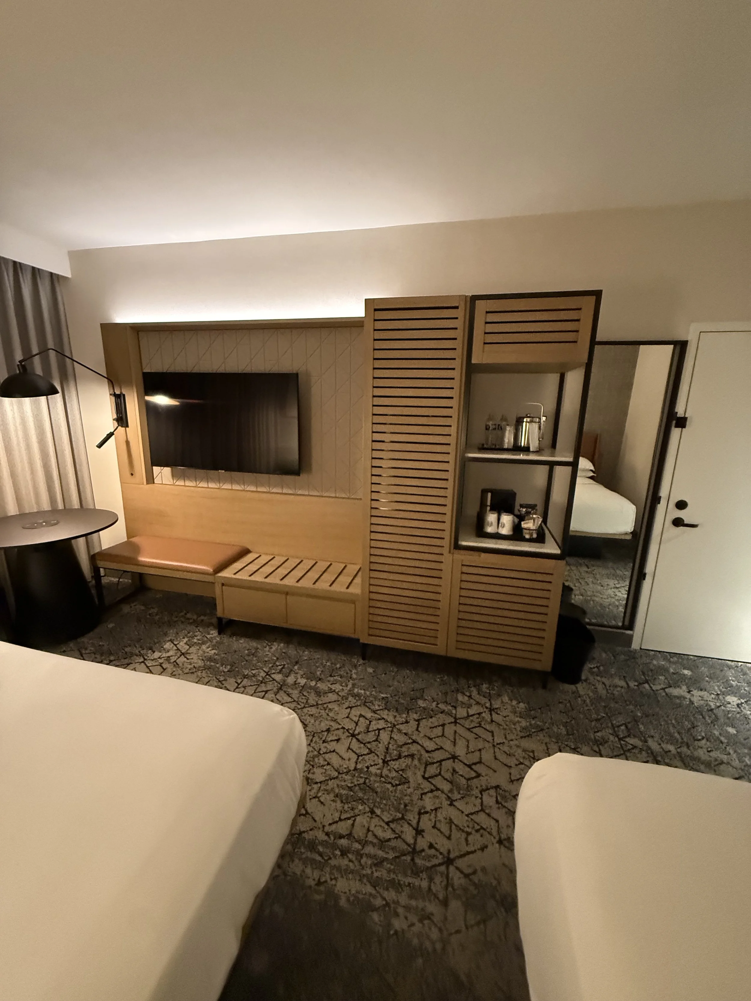 Inside a modern hotel room with two beds, a flat-screen TV mounted on a wooden partition, a small bench, a round table, a wall-mounted lamp, and a shelving unit with beverages and a mini fridge. A mirror reflects part of the room and a door is visibl