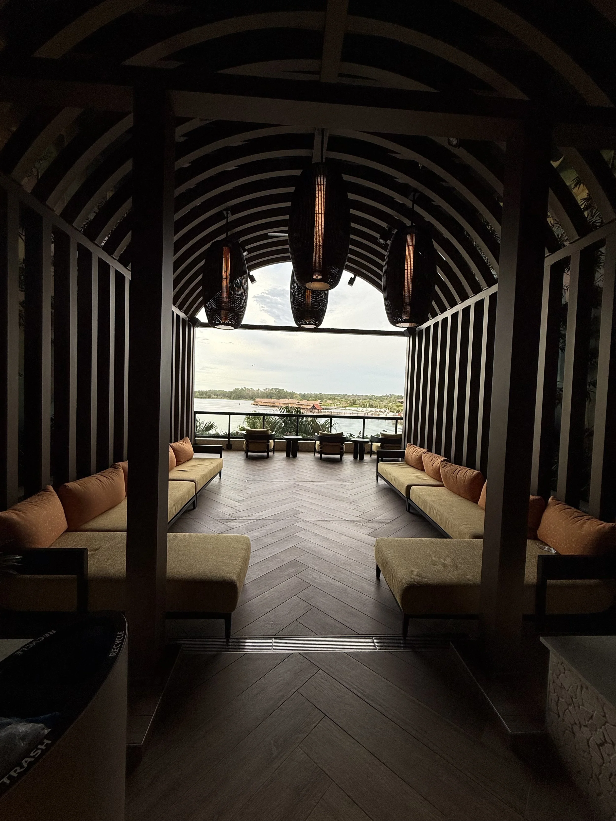 A covered outdoor seating area with beige couches and orange cushions, facing a view of water and distant landscape, with six large hanging black pendant lights inside.
