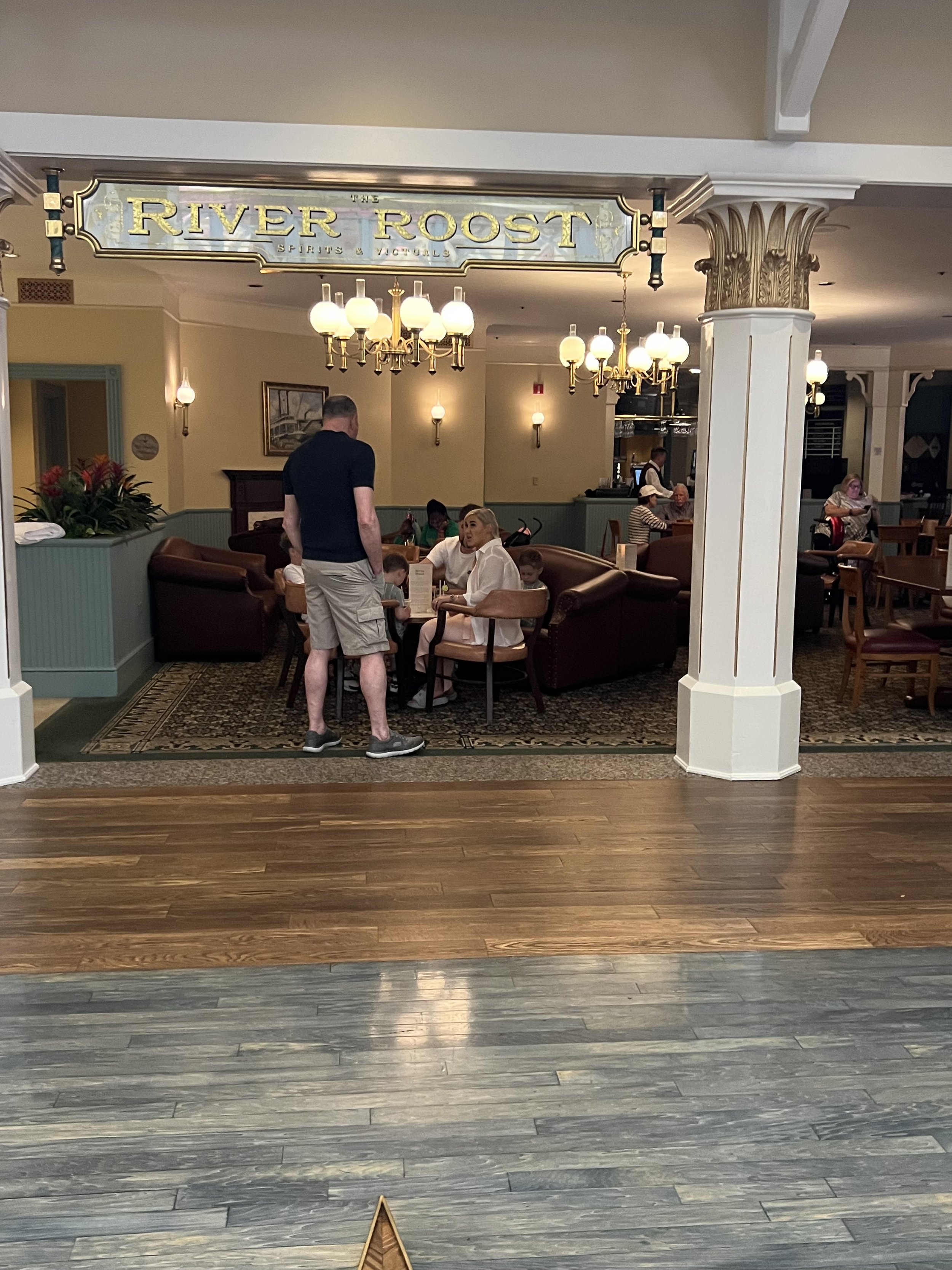 Interior of a restaurant named River Roost, with people seated at tables, a man standing, and decorative lighting fixtures.