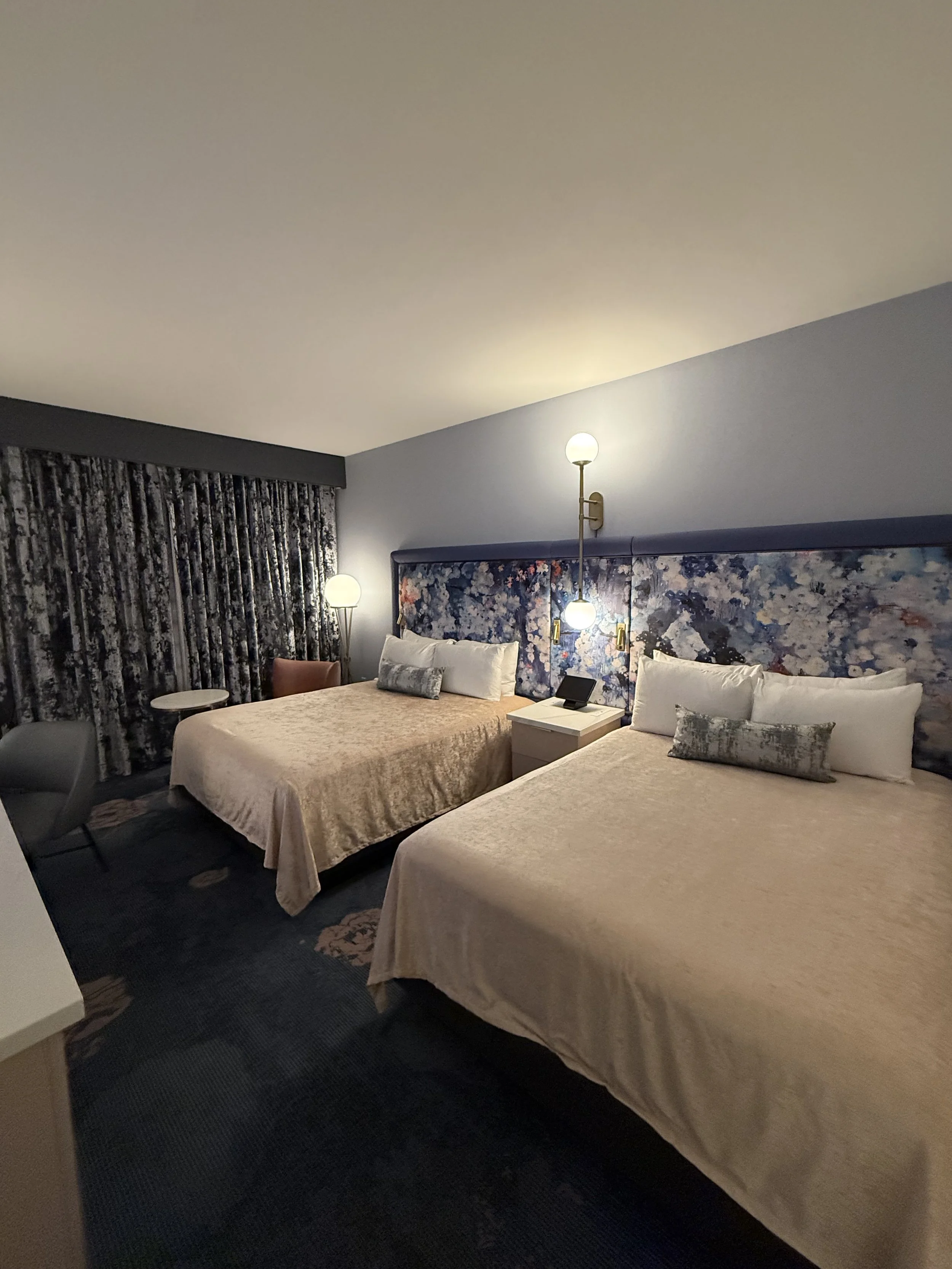 A hotel room with two beds, each with beige bedspreads and gray accent pillows. A floral headboard runs behind both beds. There are two lamps on either side of the room, one behind the bed on the left and a hanging lamp between the beds. A bedside ta