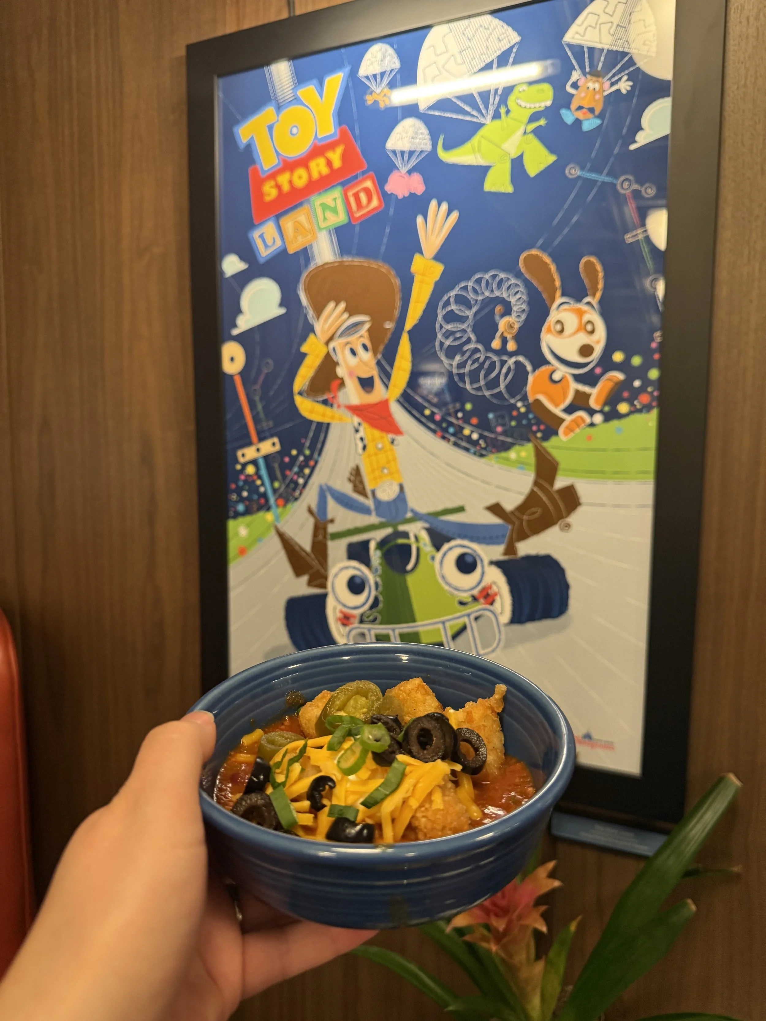 Person holding a bowl of loaded nachos topped with cheese, black olives, and green onions in front of a Toy Story Land poster featuring Woody and Rex.