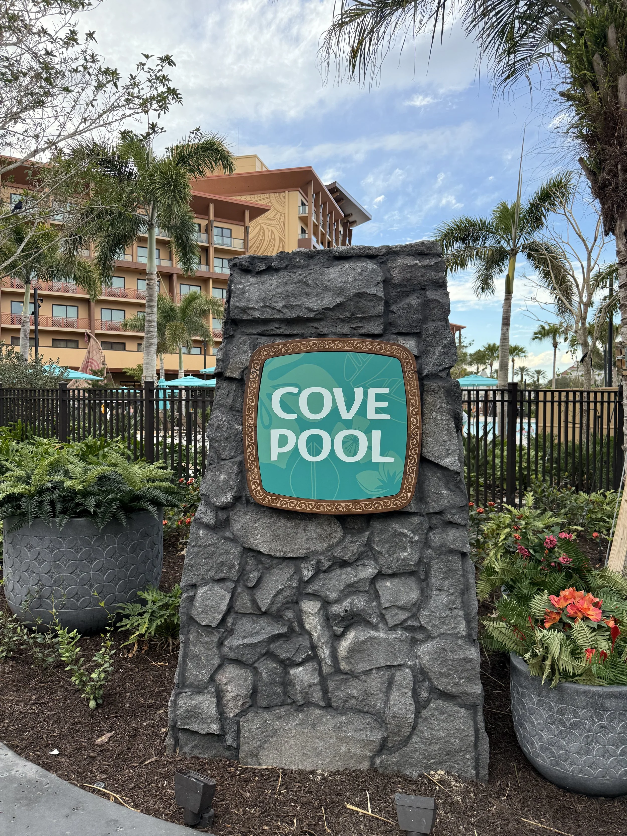 Sign for Cove Pool with a stone background and the resort building in the background, surrounded by trees and potted plants.