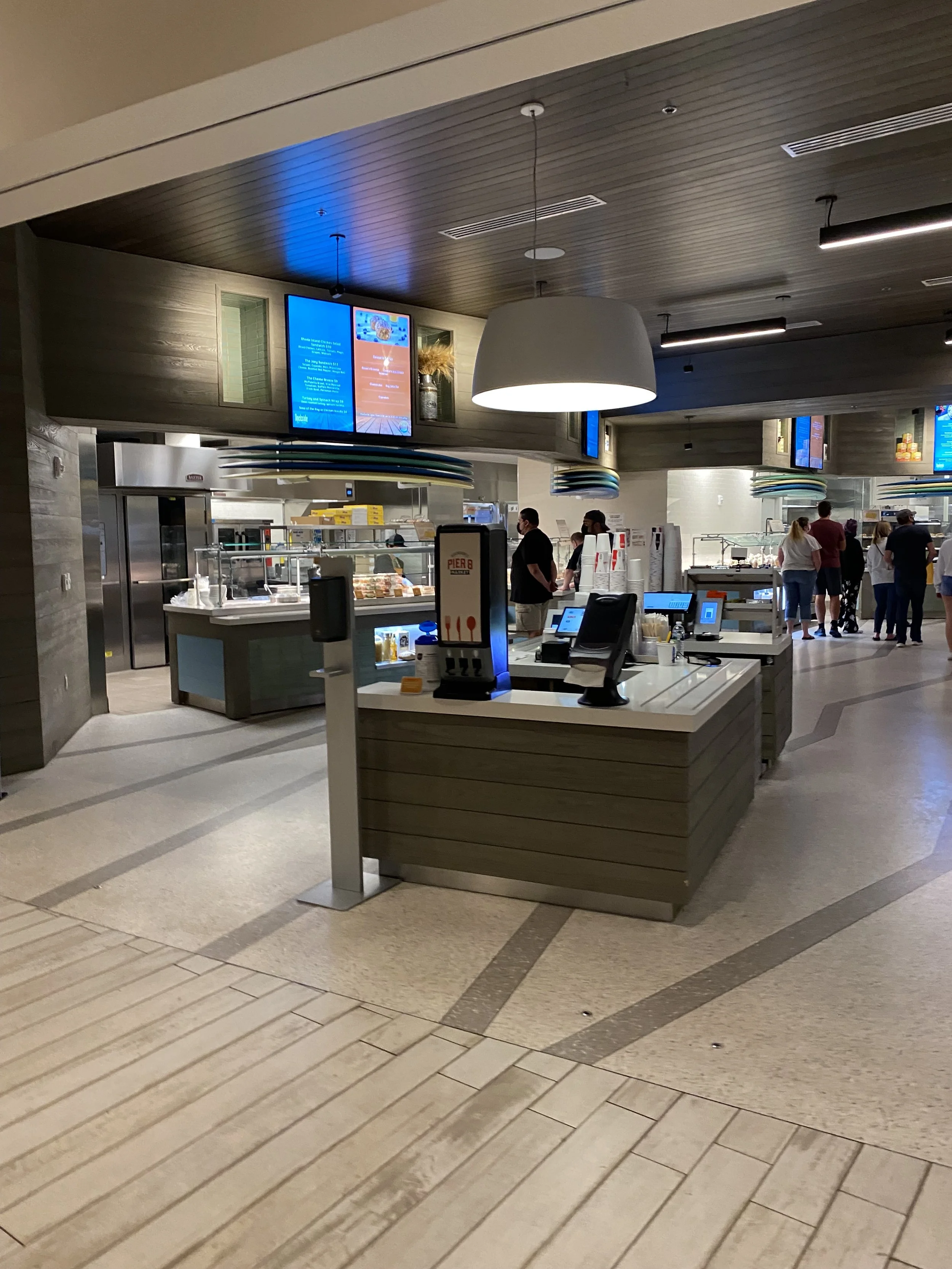 Indoor food court or cafeteria with a serving counter, digital menu screens, and a group of people in line or waiting nearby.