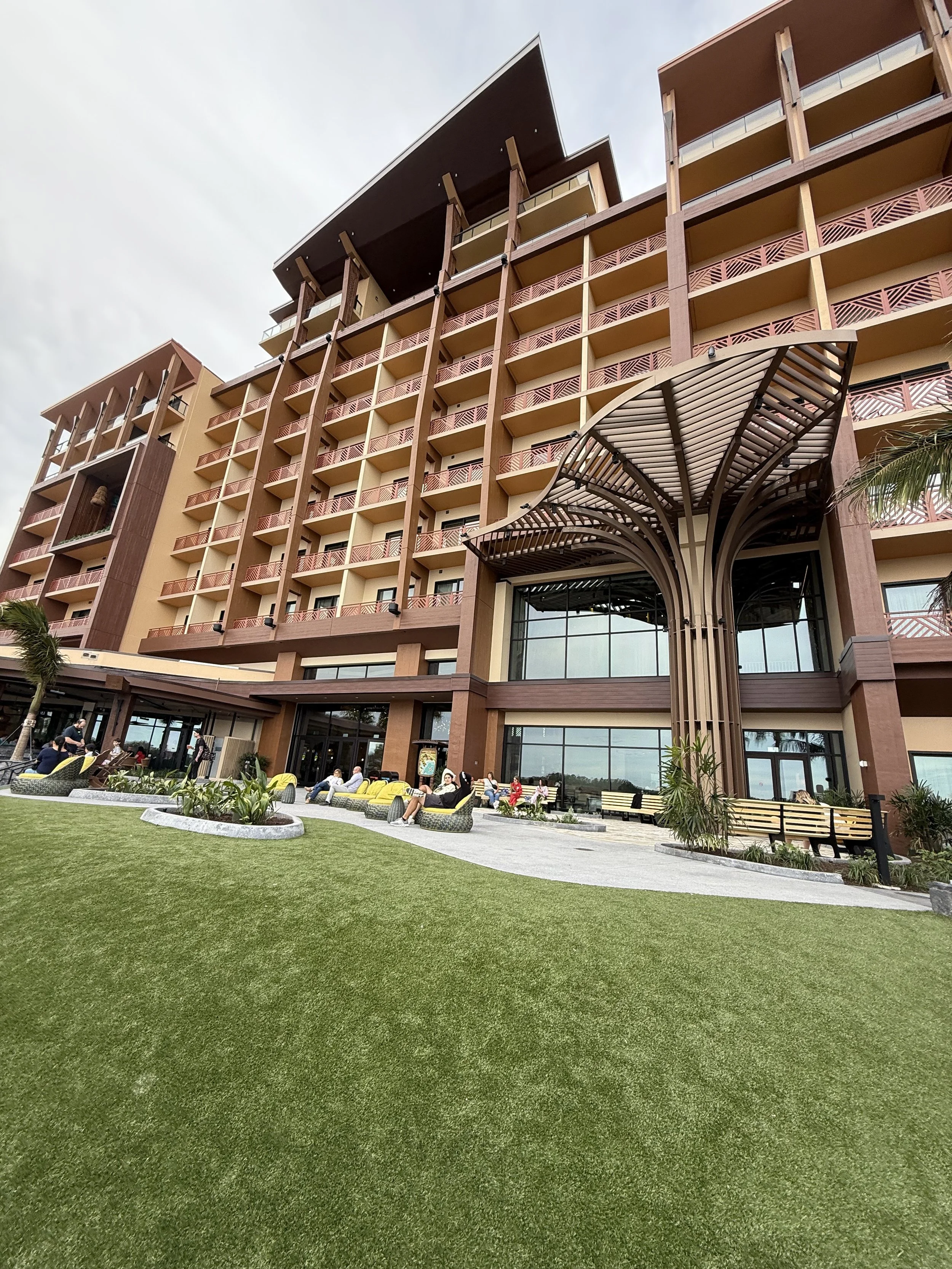A modern hotel with multiple floors and a decorative wooden canopy at the entrance. People are sitting on benches and chairs on the lawn outside, with palm trees and potted plants nearby.