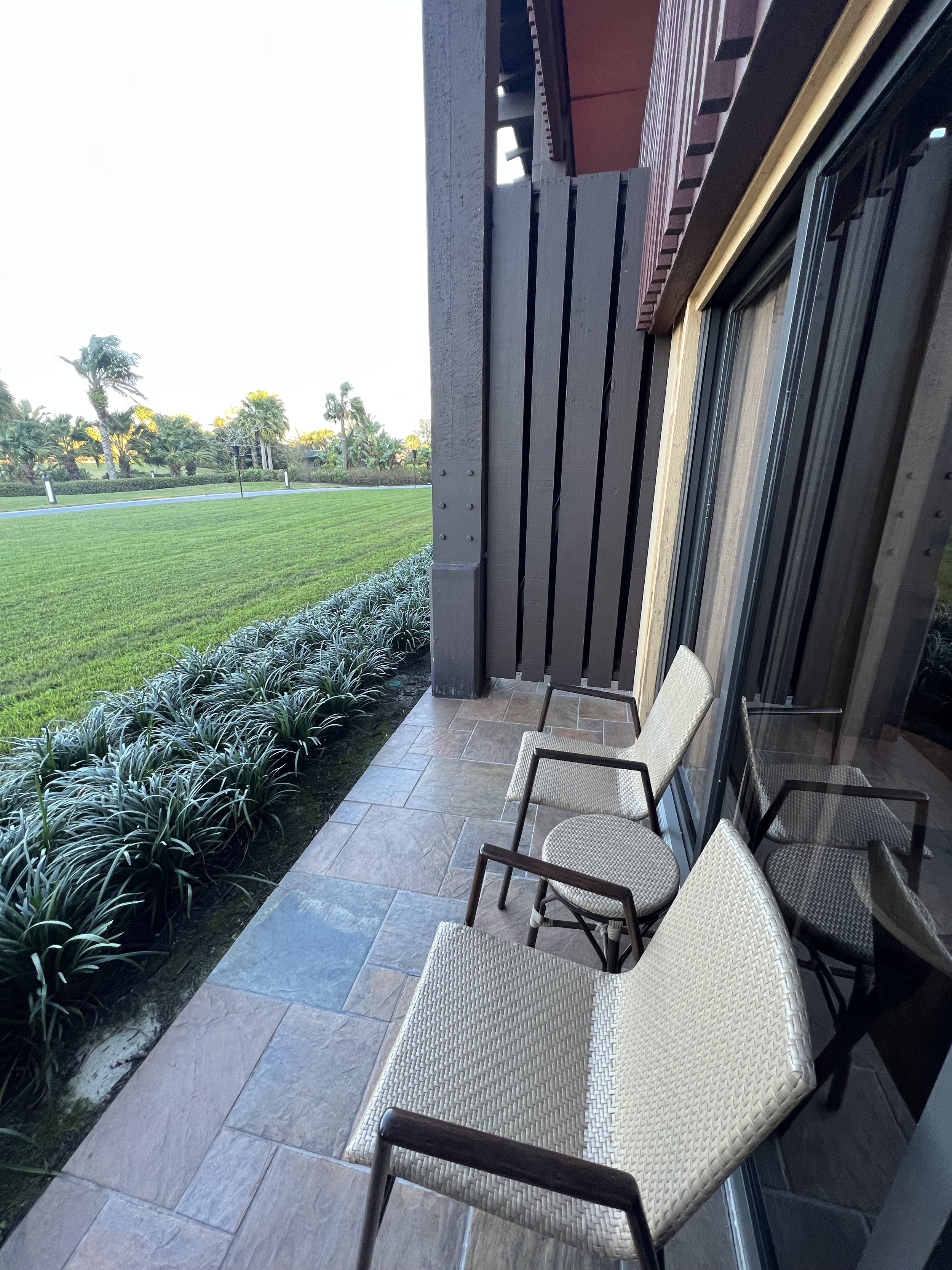 Patio with two beige chairs with black wooden armrests, a doormat, and a glass door leading to a lush green lawn and trees in the background.