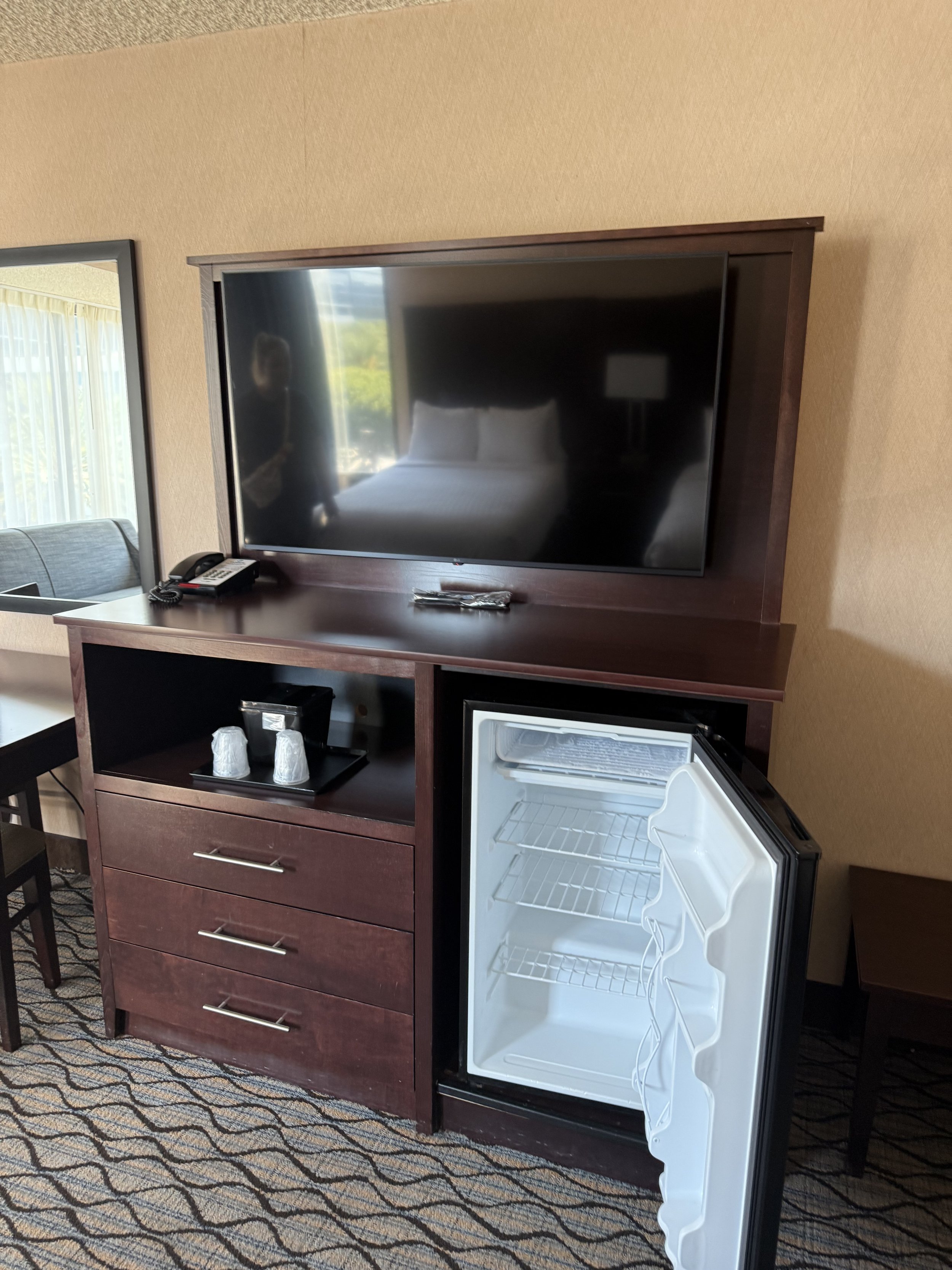 Hotel room mini fridge open beside wooden cabinet with drawers, television on top, and a reflection of the room in the TV screen.