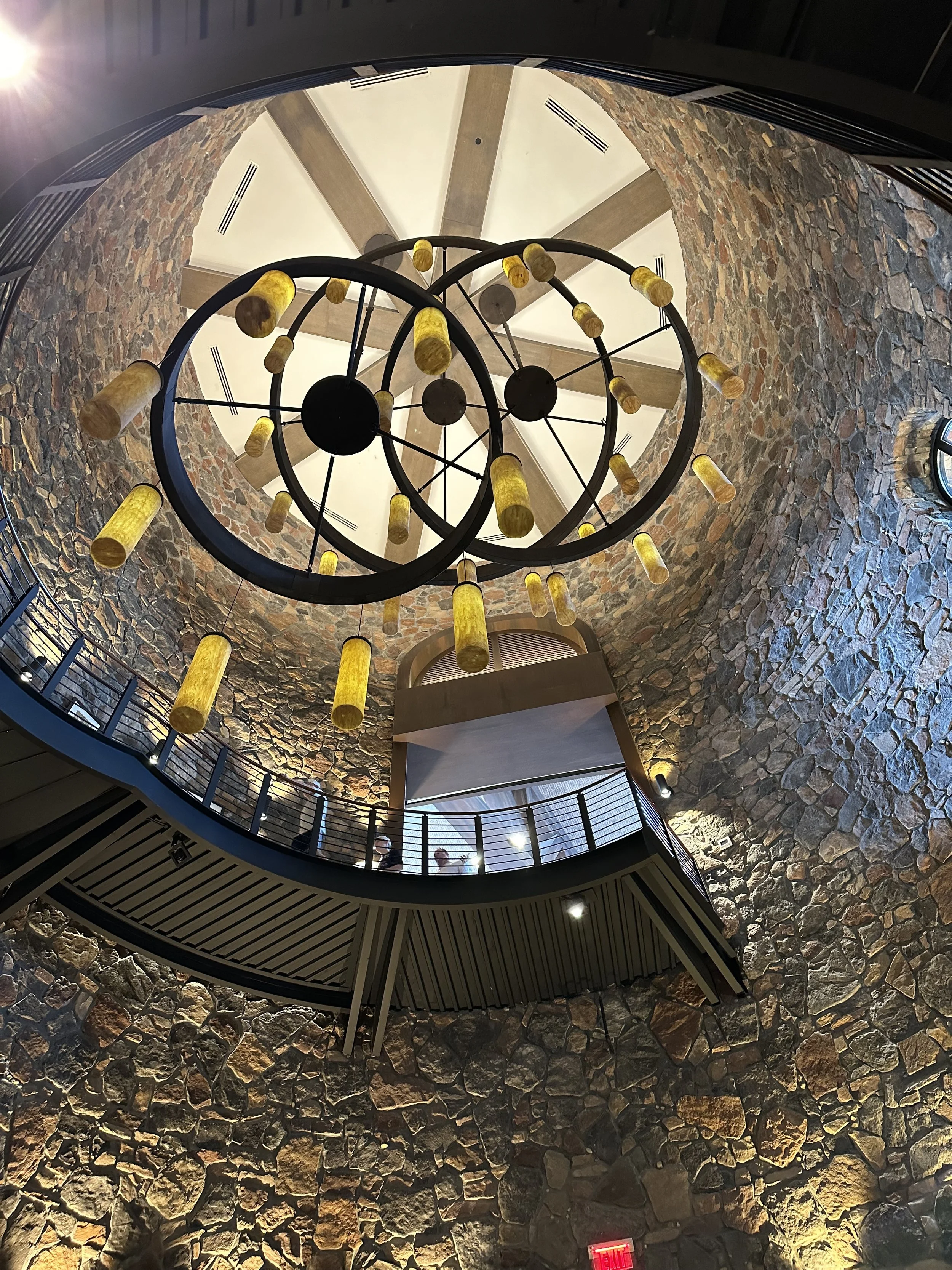 Interior view of a building with a tall stone wall, a spiral staircase, and a modern chandelier with yellow lights hanging from the ceiling.