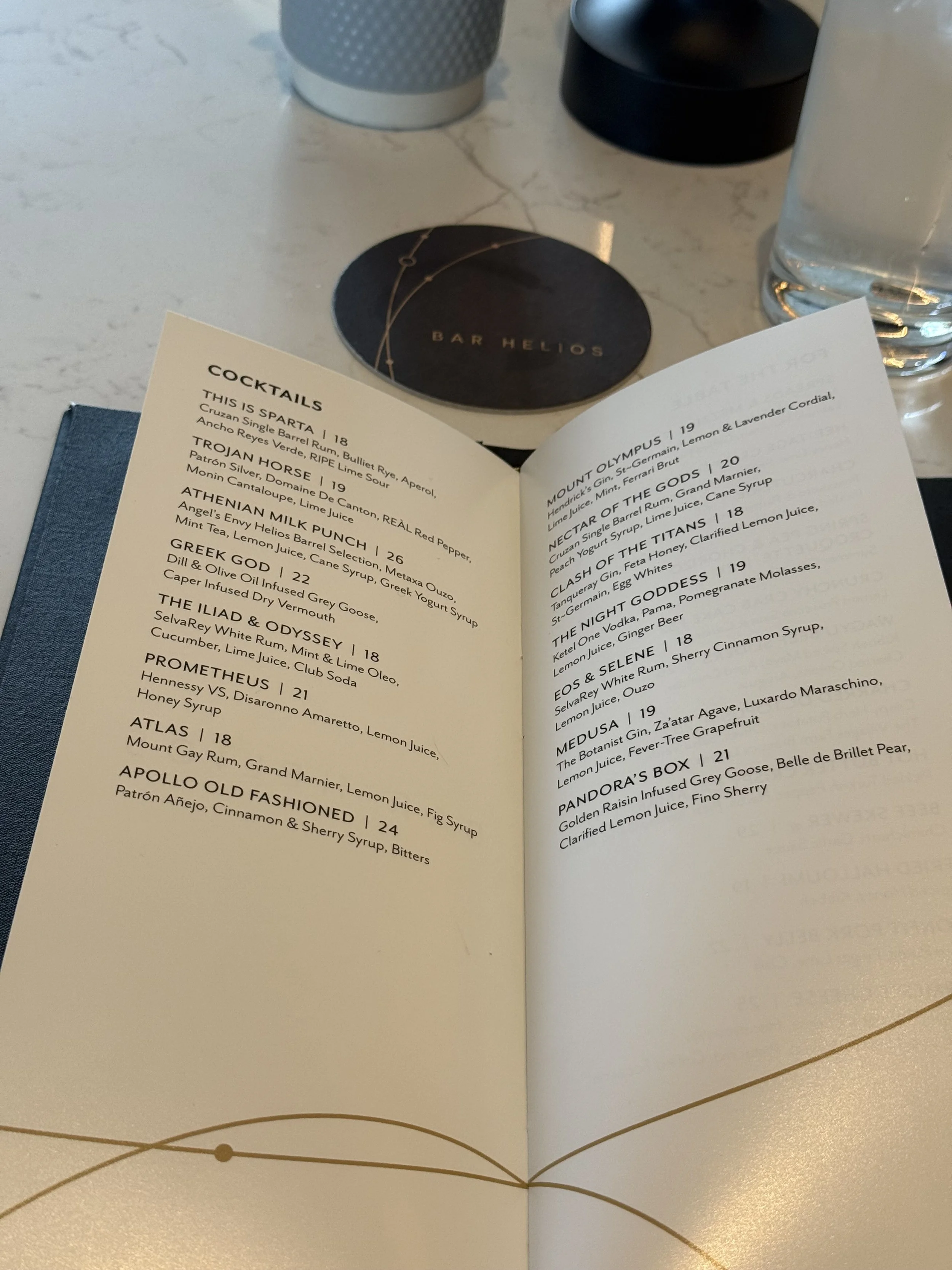 Close-up of a cocktail menu from Bar Helios featuring various drinks with their ingredients listed, placed on a table with a glass of water and coasters in the background.