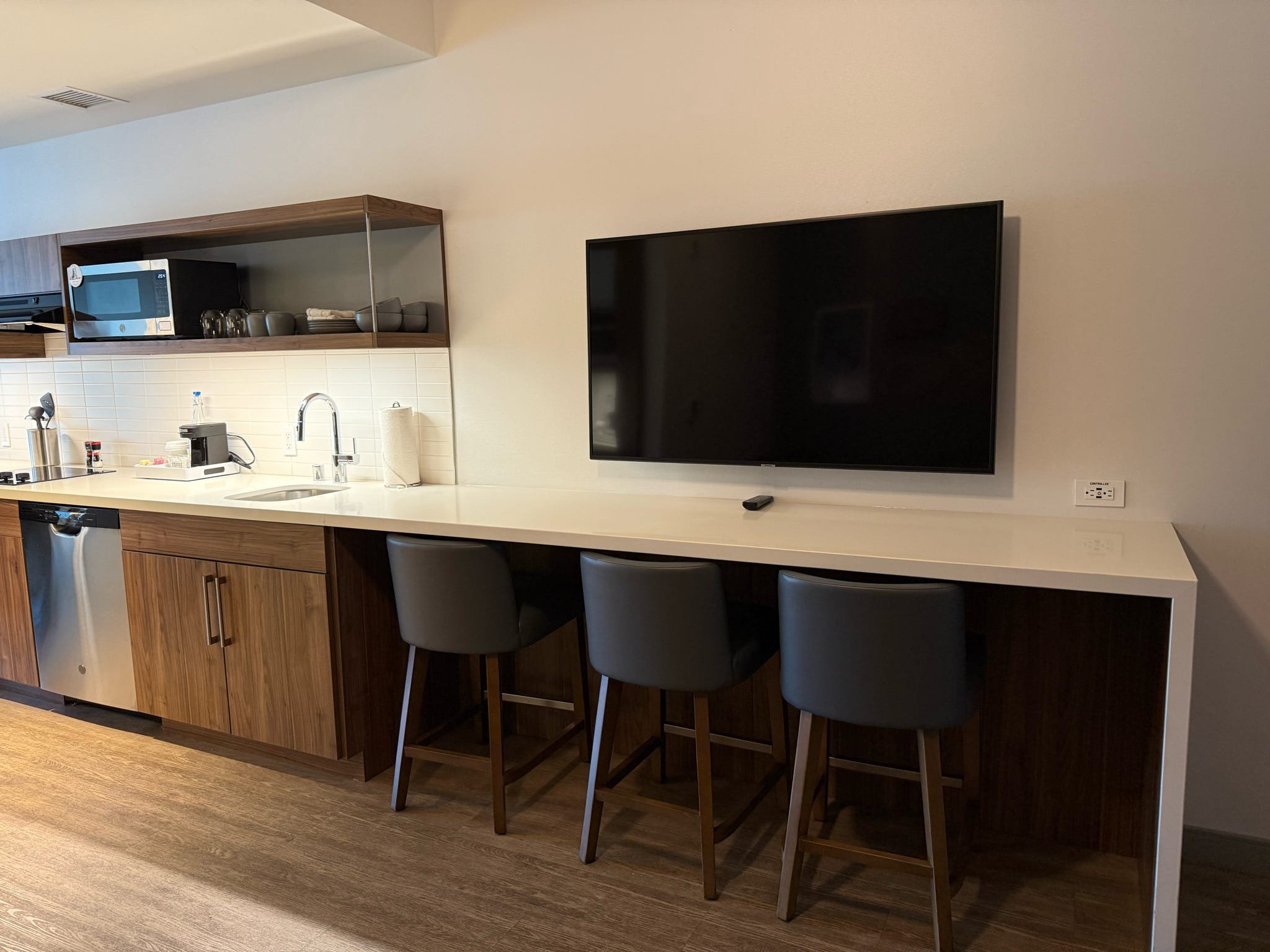 Modern kitchen area with a white countertop, three bar stools, a mounted flat-screen TV, and kitchen appliances including microwave, coffee machine, and various cups and utensils.