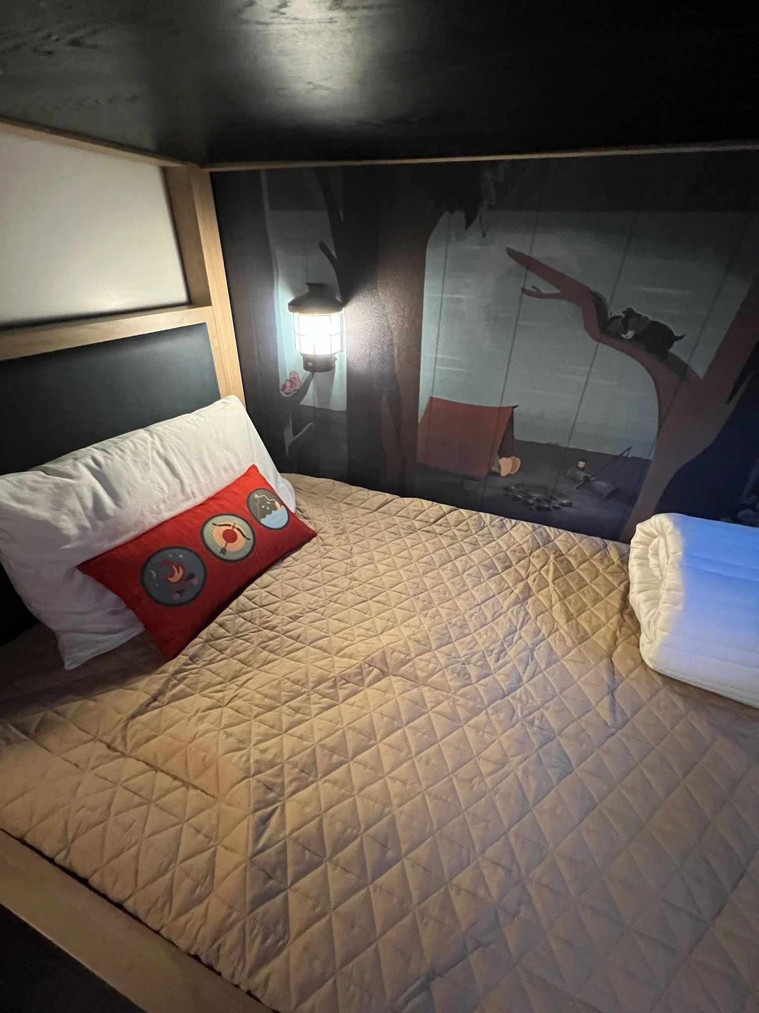 A bedroom with a bed, a beige quilted bedspread, and pillows. One pillow has a red pillowcase with four circular icons. There is a wall-mounted lamp and a mural of a tree with a swing, a tent, and a person sitting on a branch.