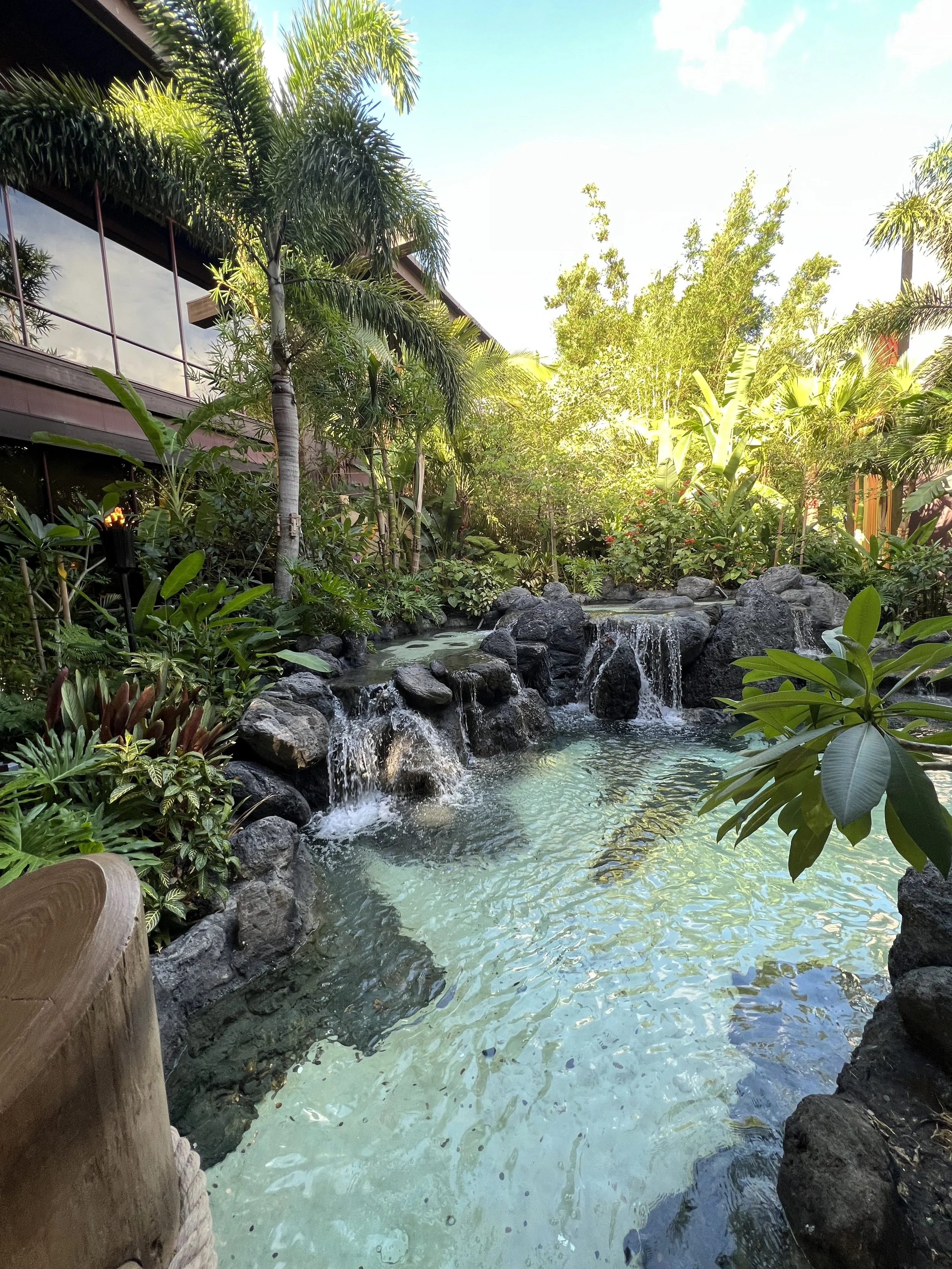 Tropical garden with rocks and small waterfalls flowing into a tranquil pool, surrounded by lush green plants and trees under a bright sky.