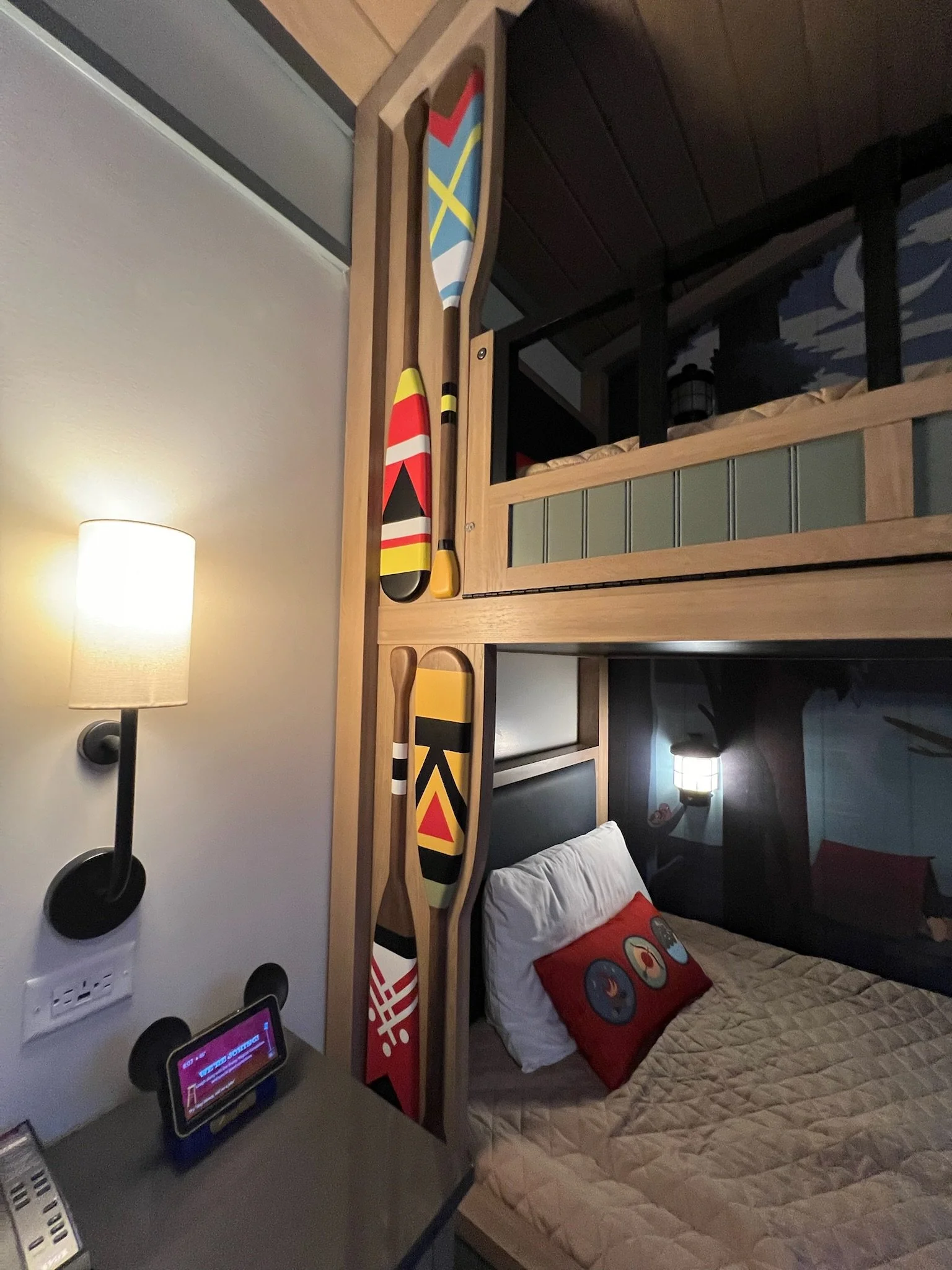 Bunk bed with colorful skis mounted on the side, a wall lamp, a smartphone on the bedside table, and a bed with a decorative pillow featuring circular designs.