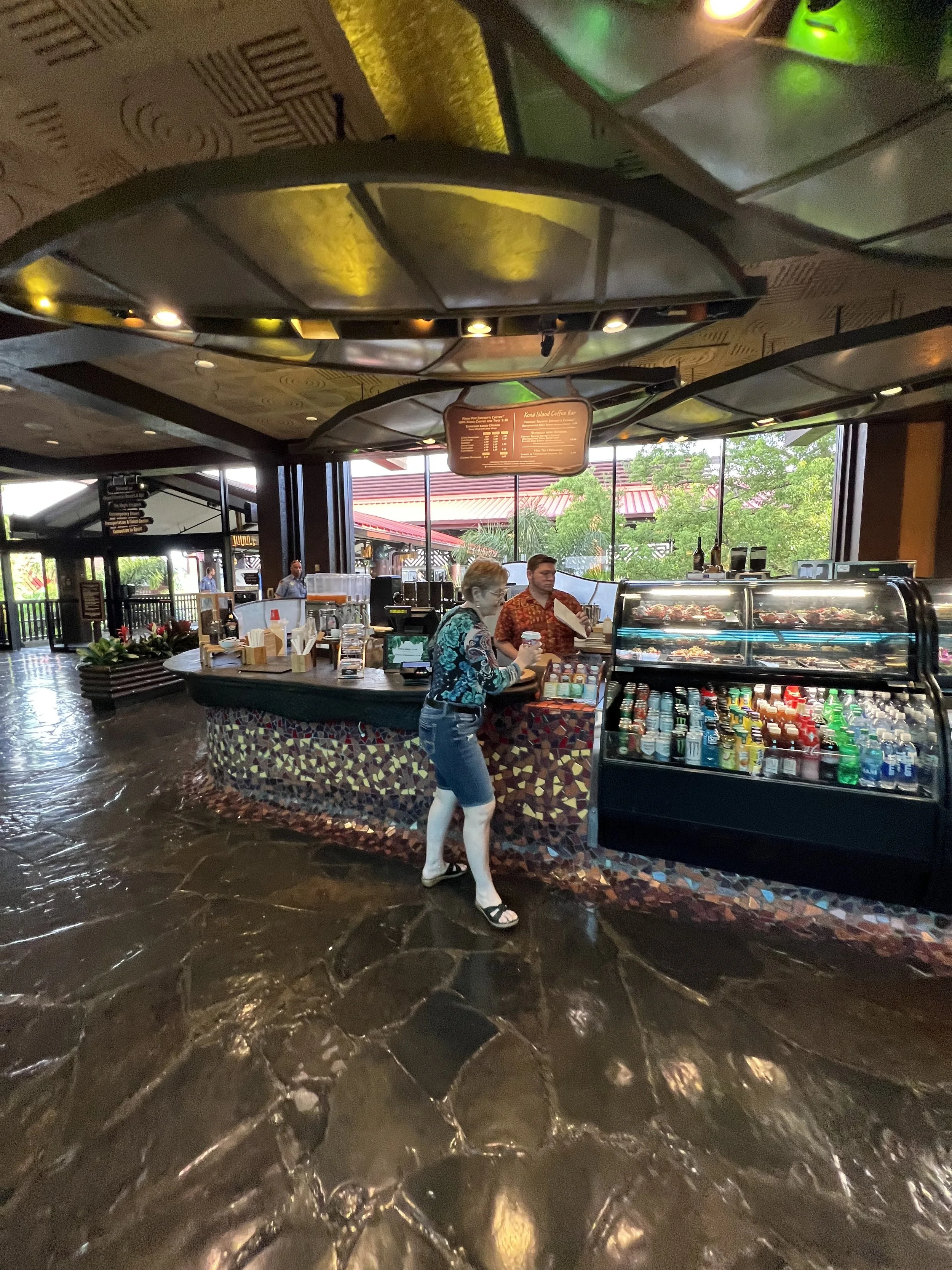 Indoor scene of a cafe or restaurant with a curved counter, a display case filled with drinks or desserts, and two staff members behind the counter. A woman is placing an order with a staff member. The ceiling has decorative panels and a digital menu