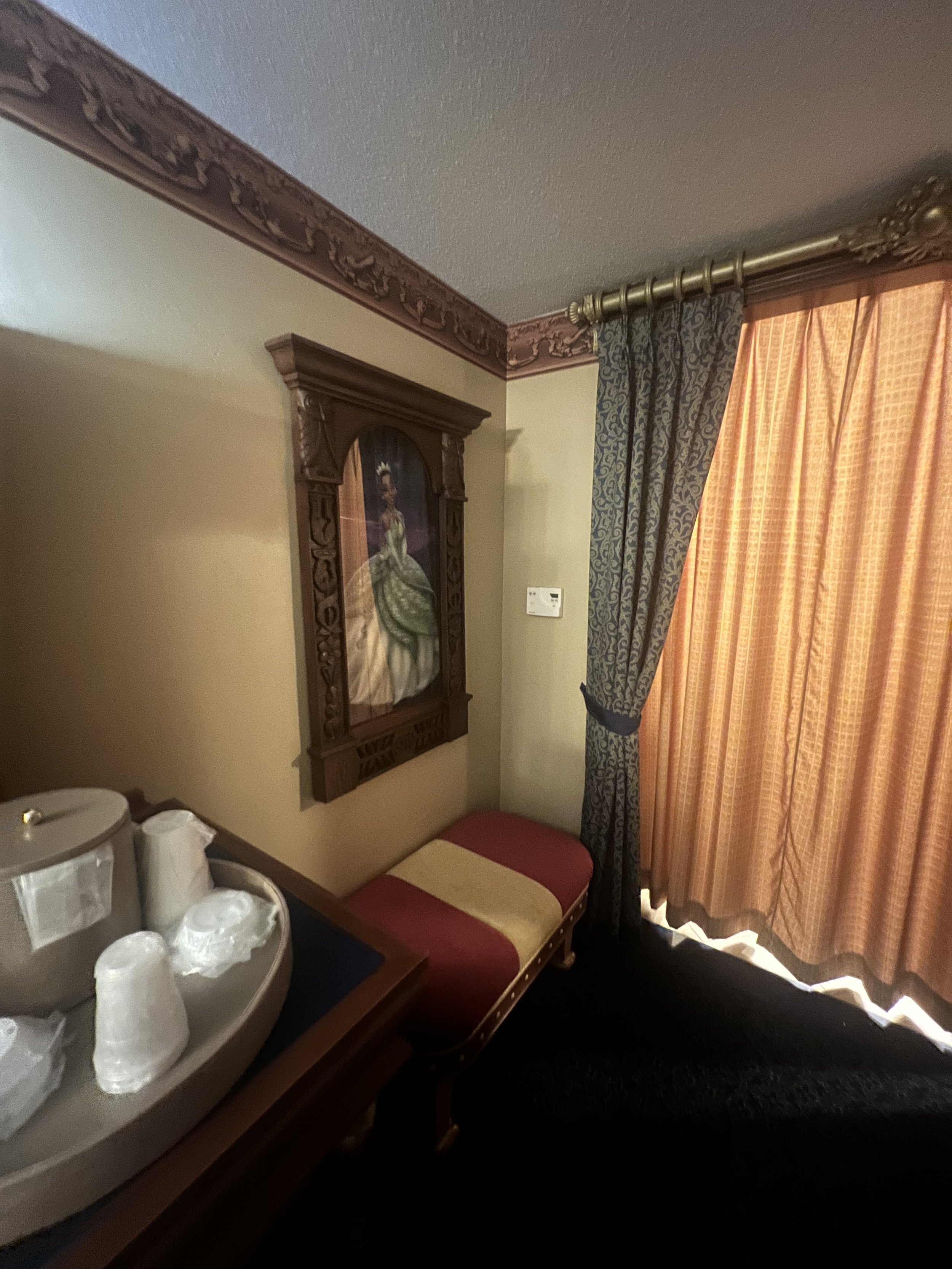 A hotel room corner with a framed painting of a woman in historic attire hanging on the wall, an ornate curtain partly covering a window, a red and beige cushioned bench, and a wooden table with cups, paper towels, and a water dispenser.