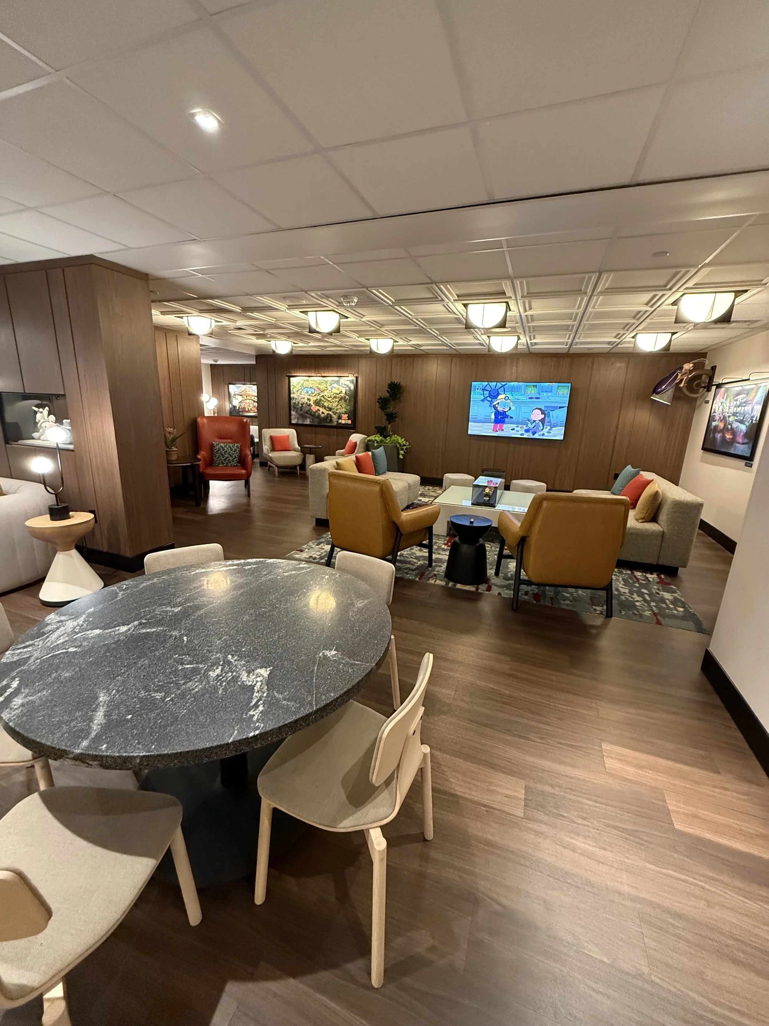 A cozy and stylish lounge area with a round marble-top table surrounded by beige chairs, and in the background, multiple sofas and armchairs arranged around a coffee table, with TV screens on the wall showing animated content. The room features wood-