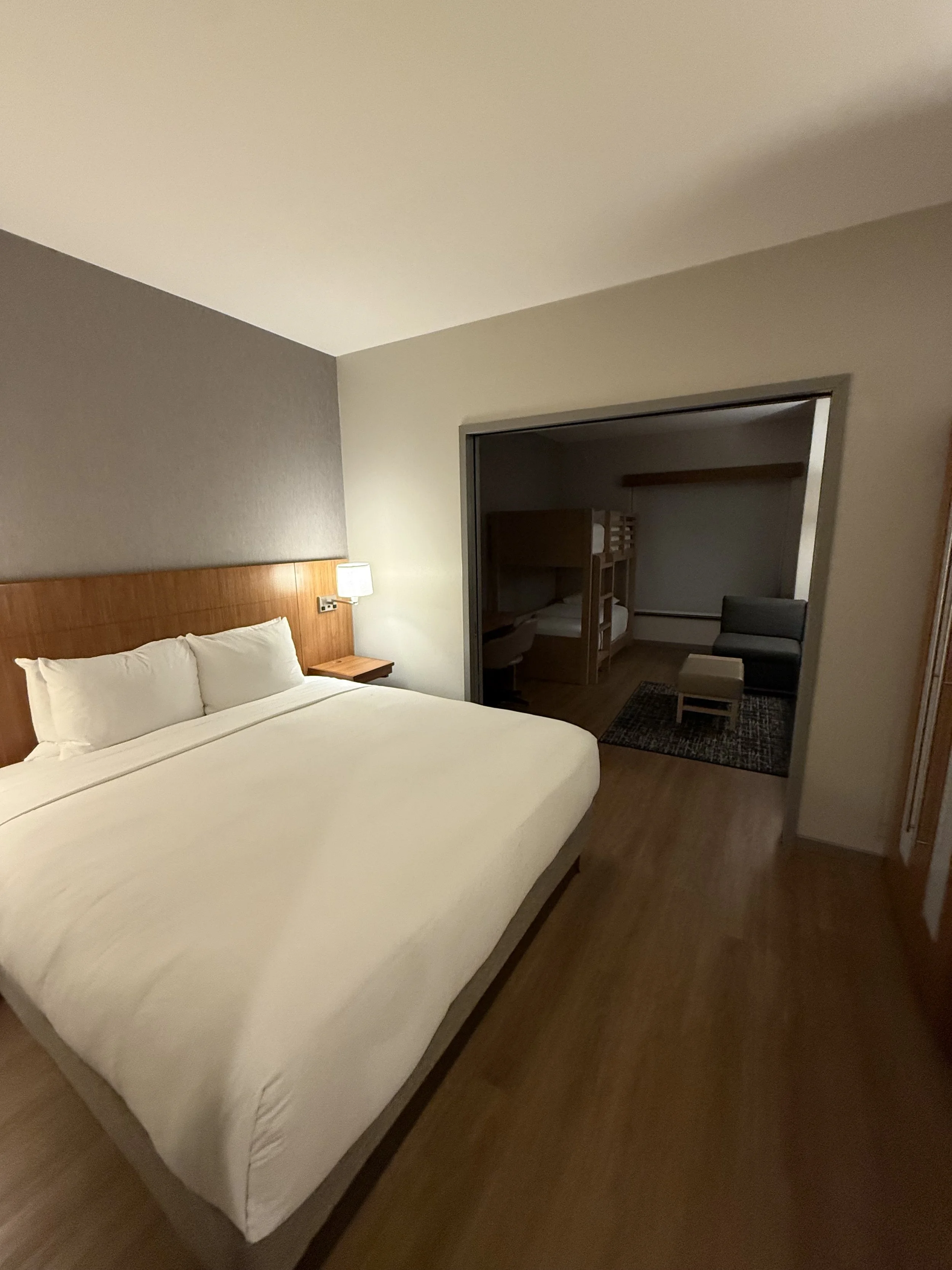 Hotel room with a large bed, bedside lamp, and a sitting area with a chair, ottoman, and bunk beds in a separate alcove.