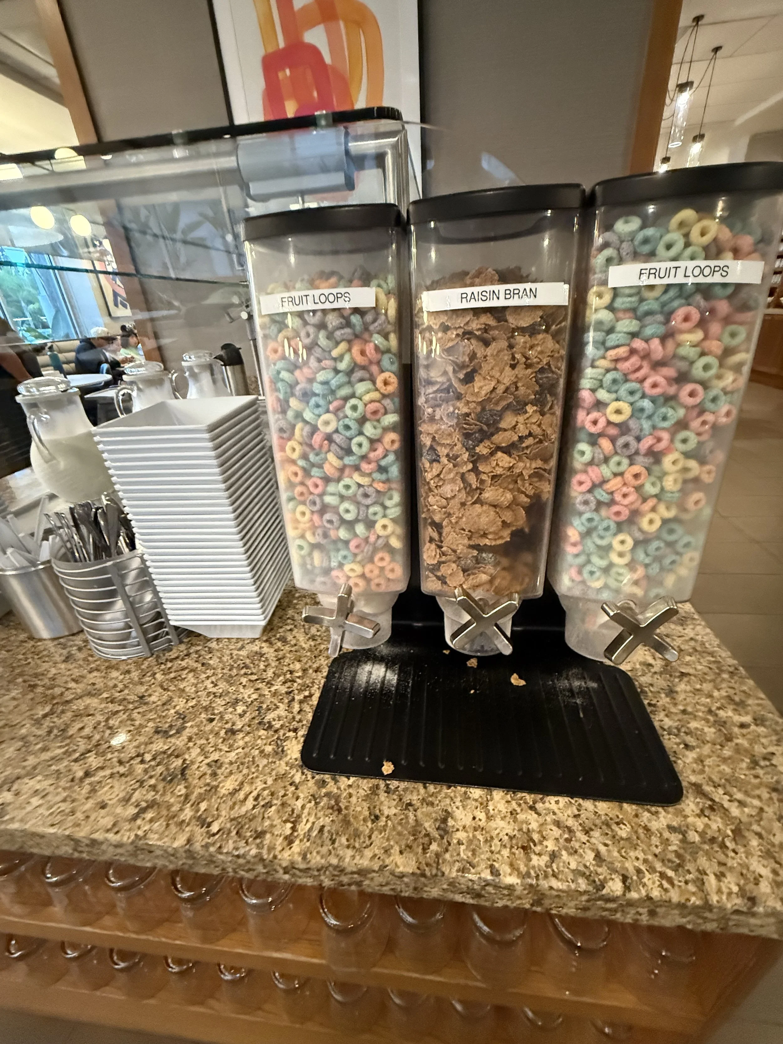 Three cereal dispensers labeled 'Fruit Loops,' 'Raisin Bran,' and 'Fruit Loops' on a granite countertop in a dining or breakfast area, with a stack of white bowls and silverware to the left.